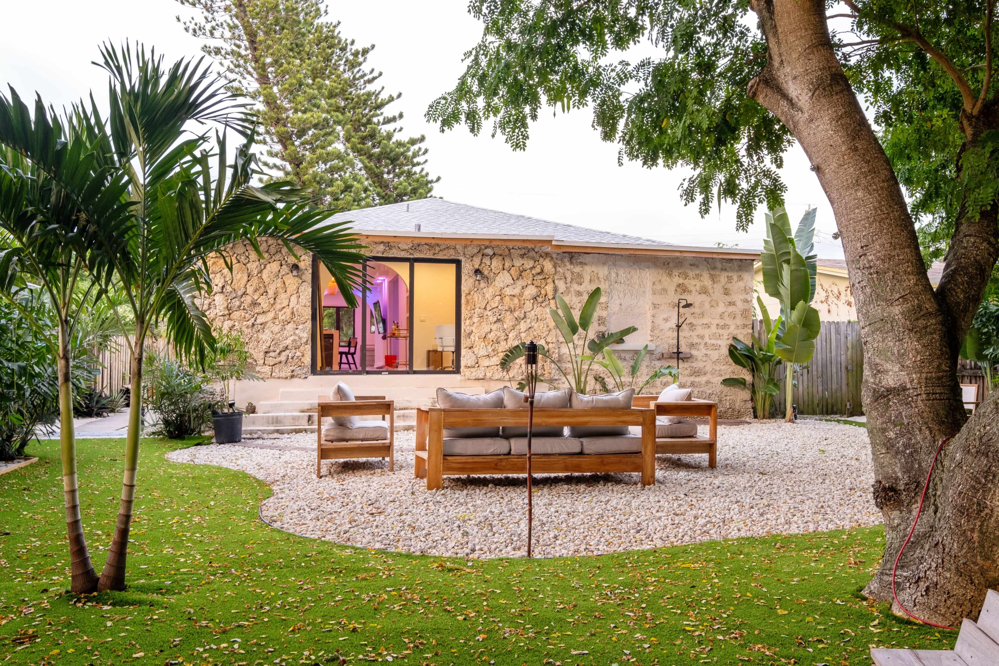 A wooden seating area is set on gravel in a landscaped yard with tropical plants, facing a stone house.