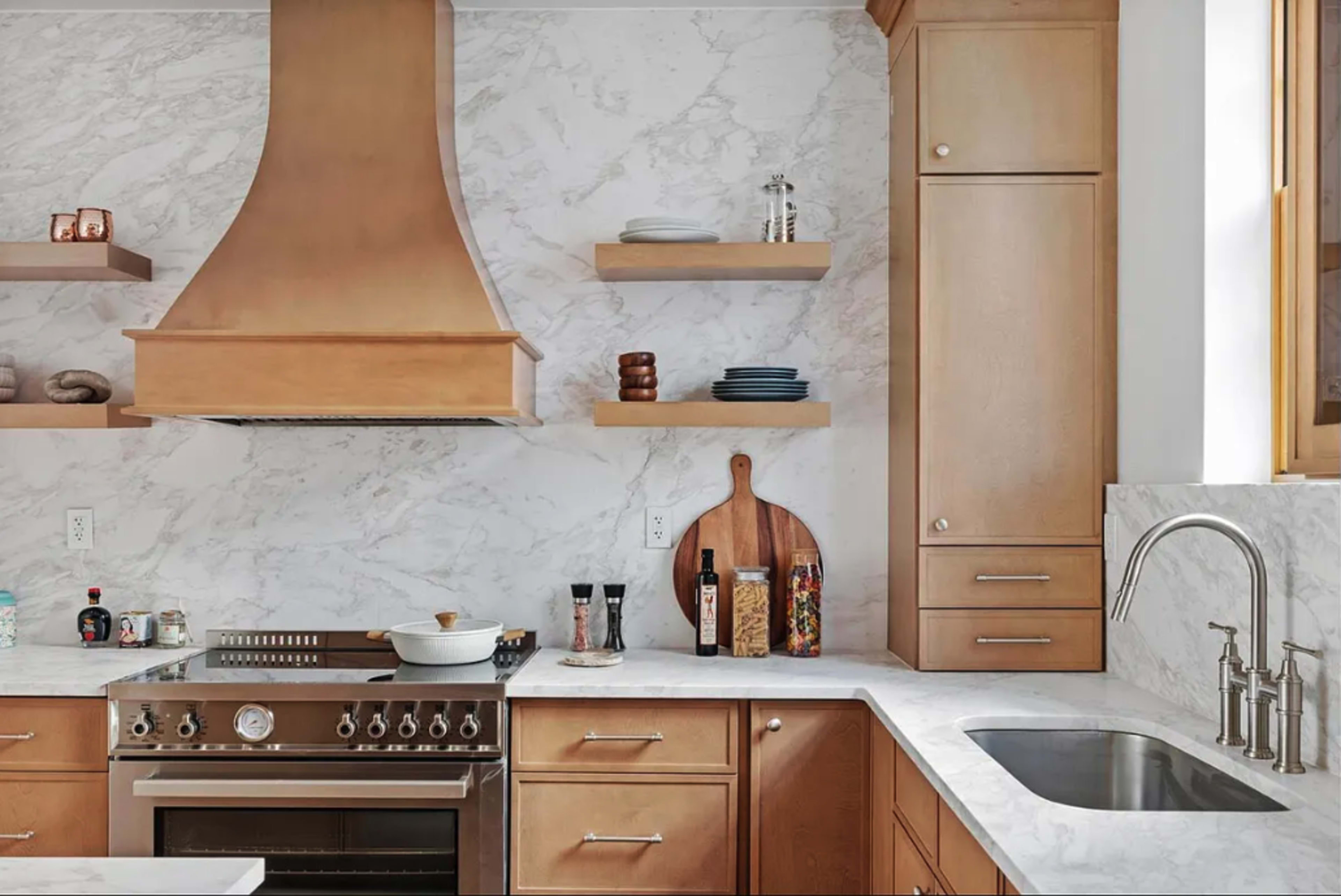 The image shows a modern kitchen featuring a marble backsplash, wooden cabinets, stainless steel appliances, and a clean countertop with a bowl and cooking utensils.
