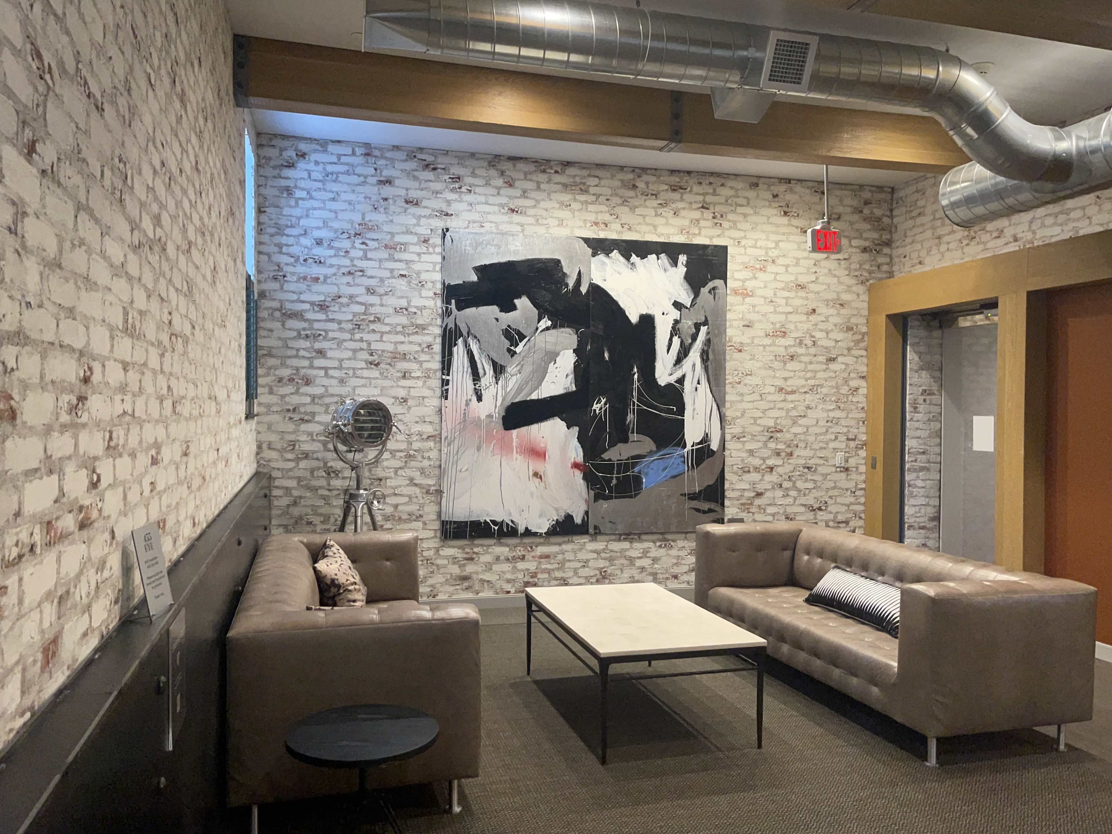 A modern lounge area with two brown leather sofas facing a coffee table, a large abstract painting on the wall, and exposed brick walls.