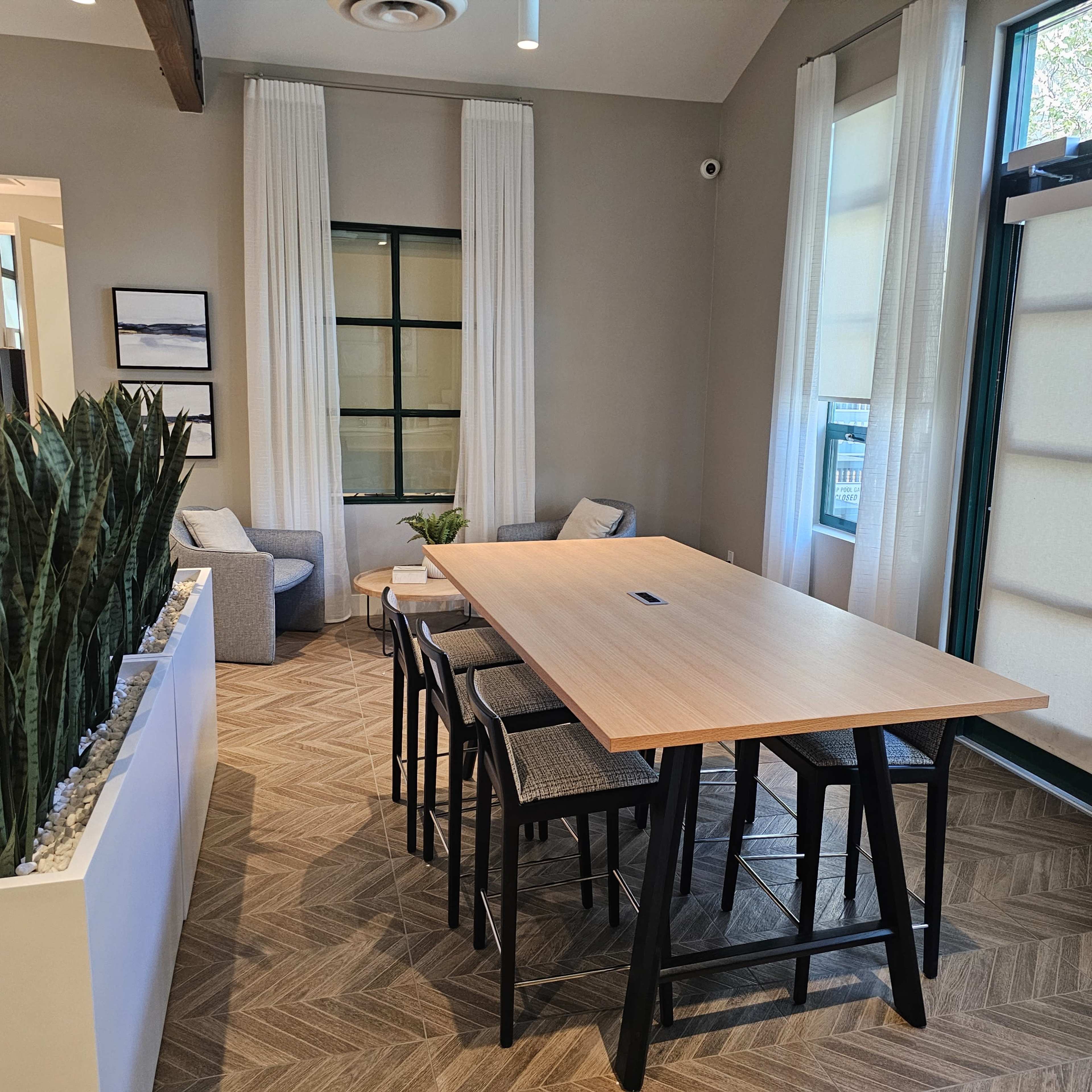 A modern, well-lit room featuring a long wooden table with high chairs, surrounded by potted plants and comfortable seating areas.