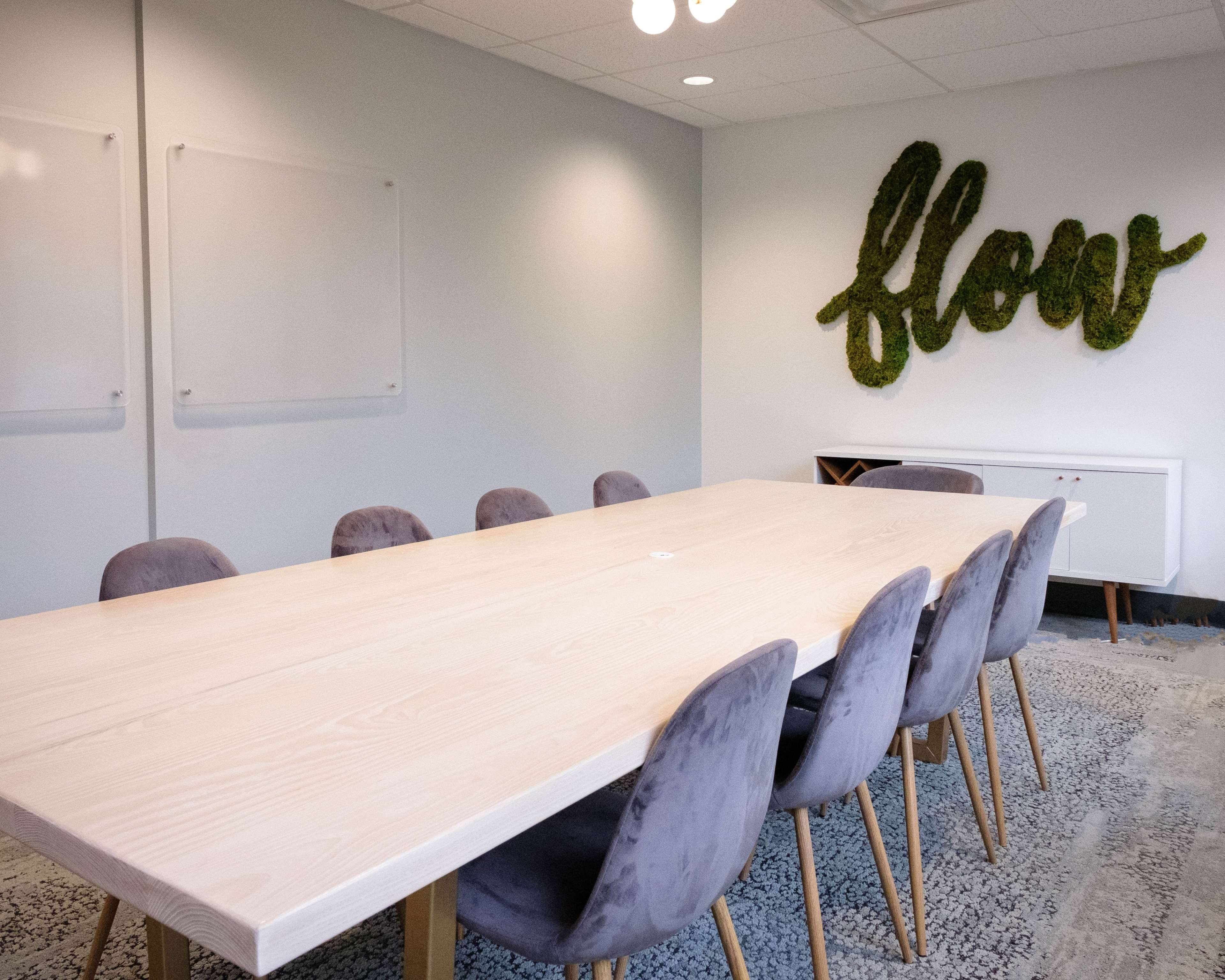 A long wooden table with eight upholstered chairs is positioned in a meeting room featuring a wall with the word "flow" made of greenery.