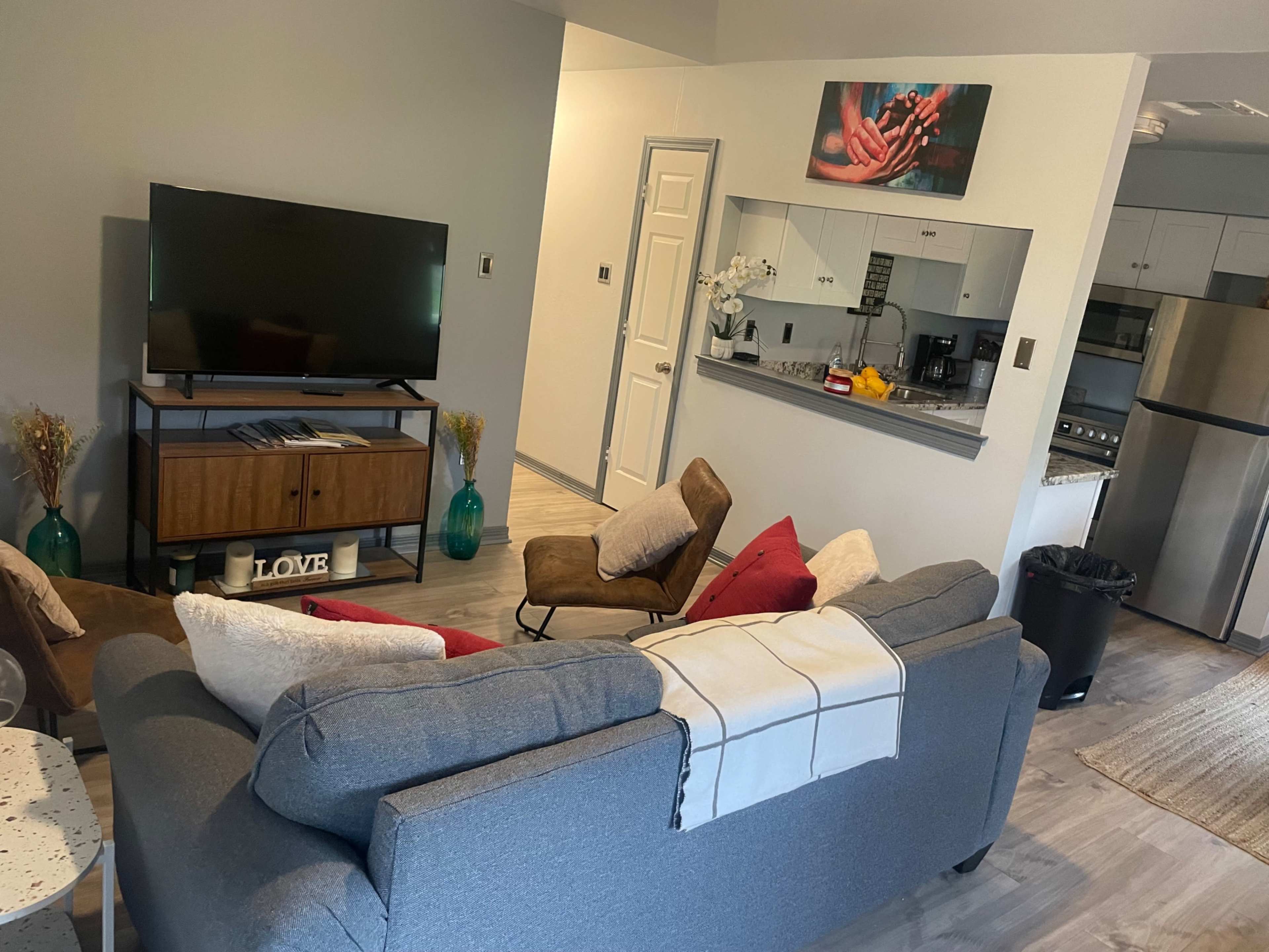 A modern living area features a gray sofa with red and beige cushions, a television on a wooden stand, and an open kitchen in the background.