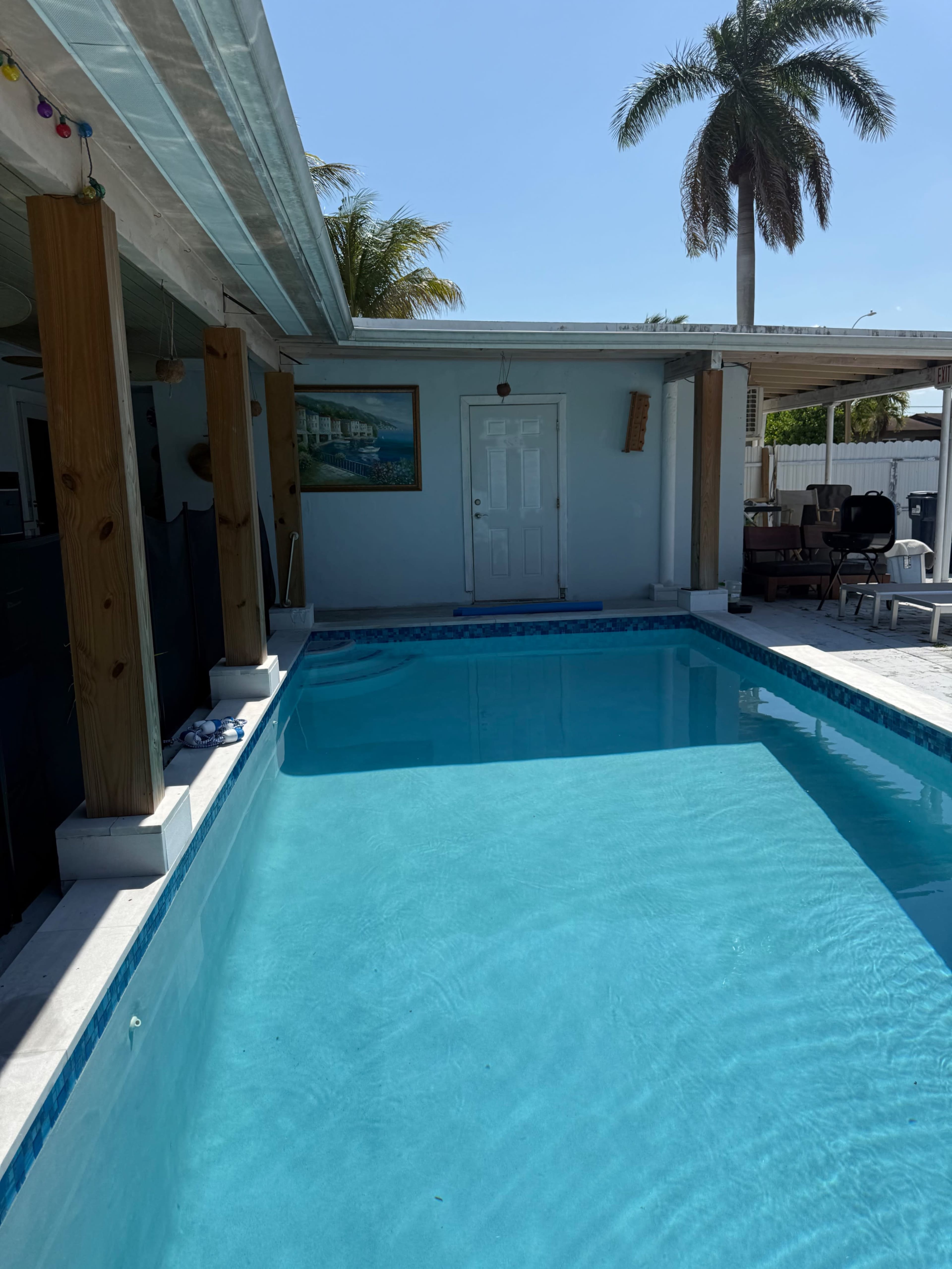 The image shows a clear blue swimming pool adjacent to a house with palm trees in the background.