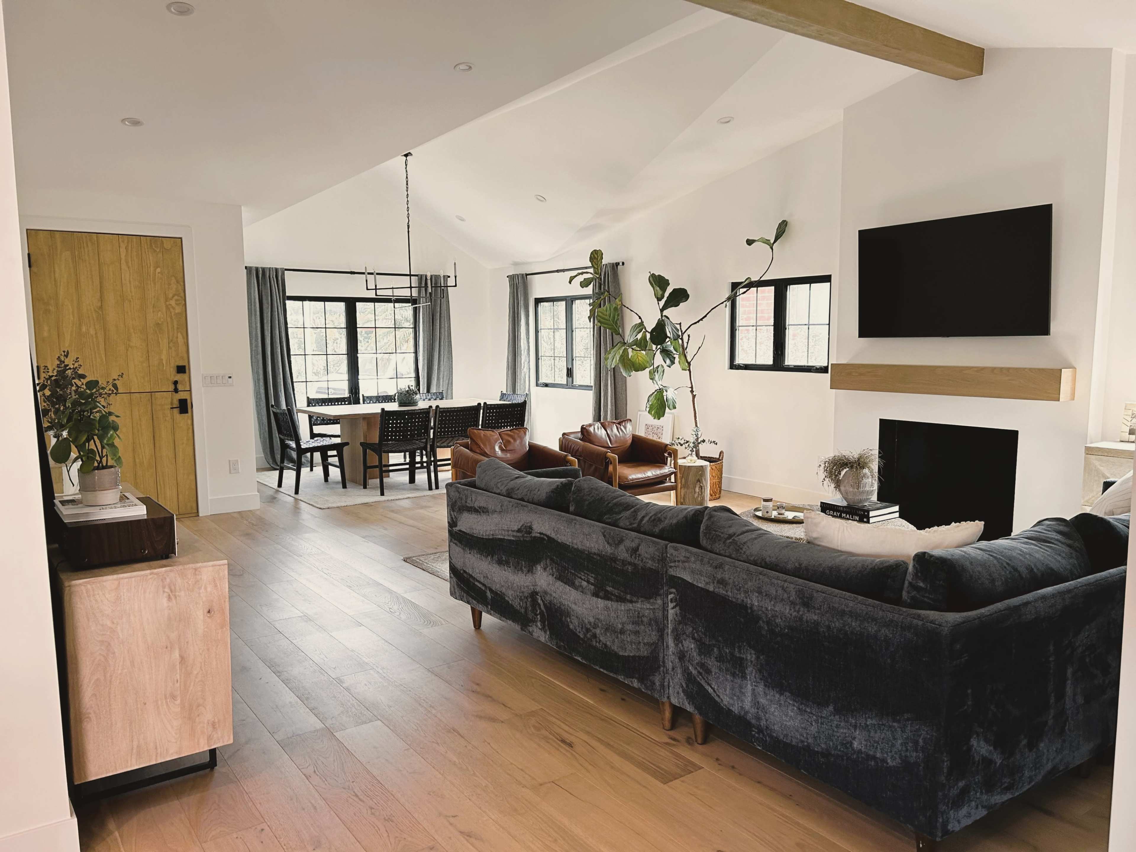 A spacious living area showcases a dark gray sofa, leather chairs, and a dining space with a table set for six, all under a high ceiling with natural light.