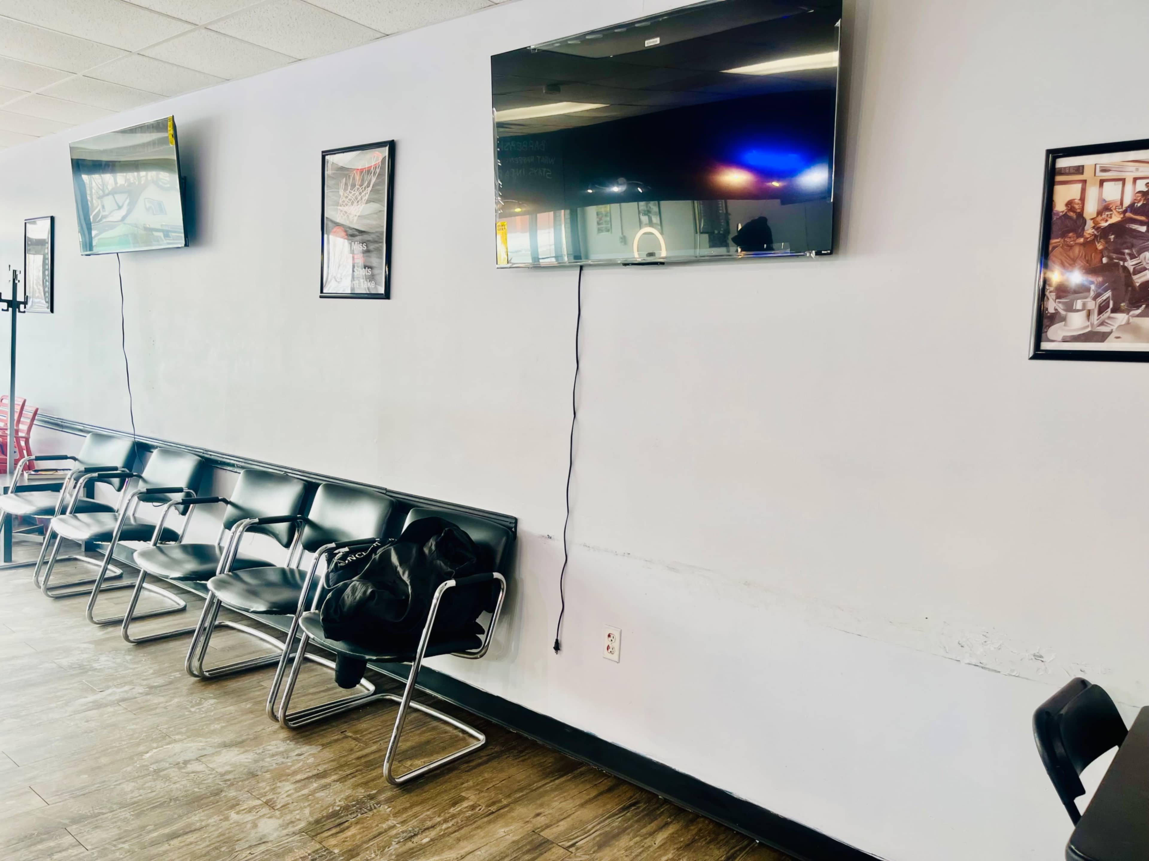 The image shows a waiting area with several black chairs, two wall-mounted televisions, and framed pictures on the walls.