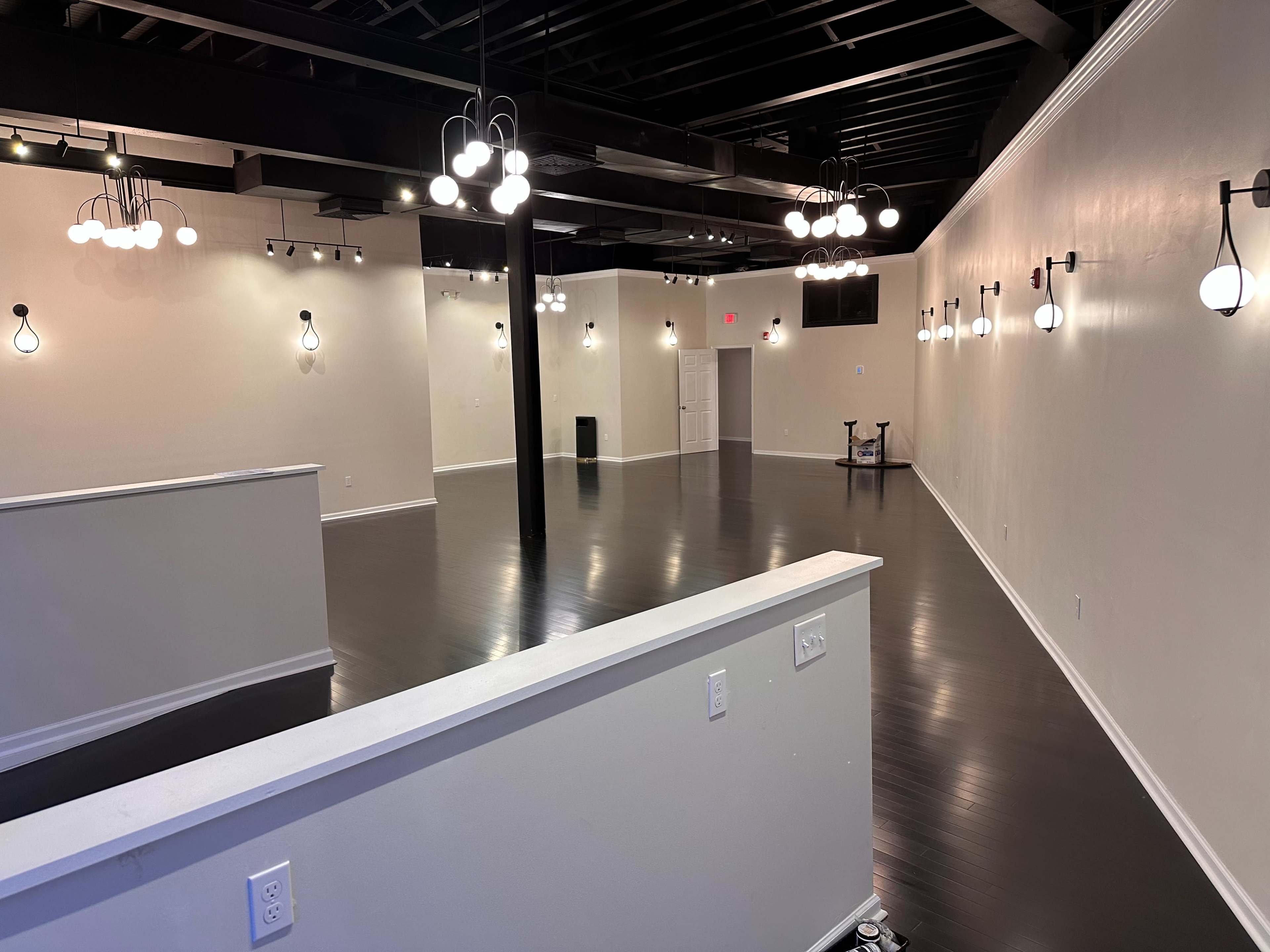 The image shows a spacious, empty interior with polished dark flooring and various hanging light fixtures on the walls and ceiling.