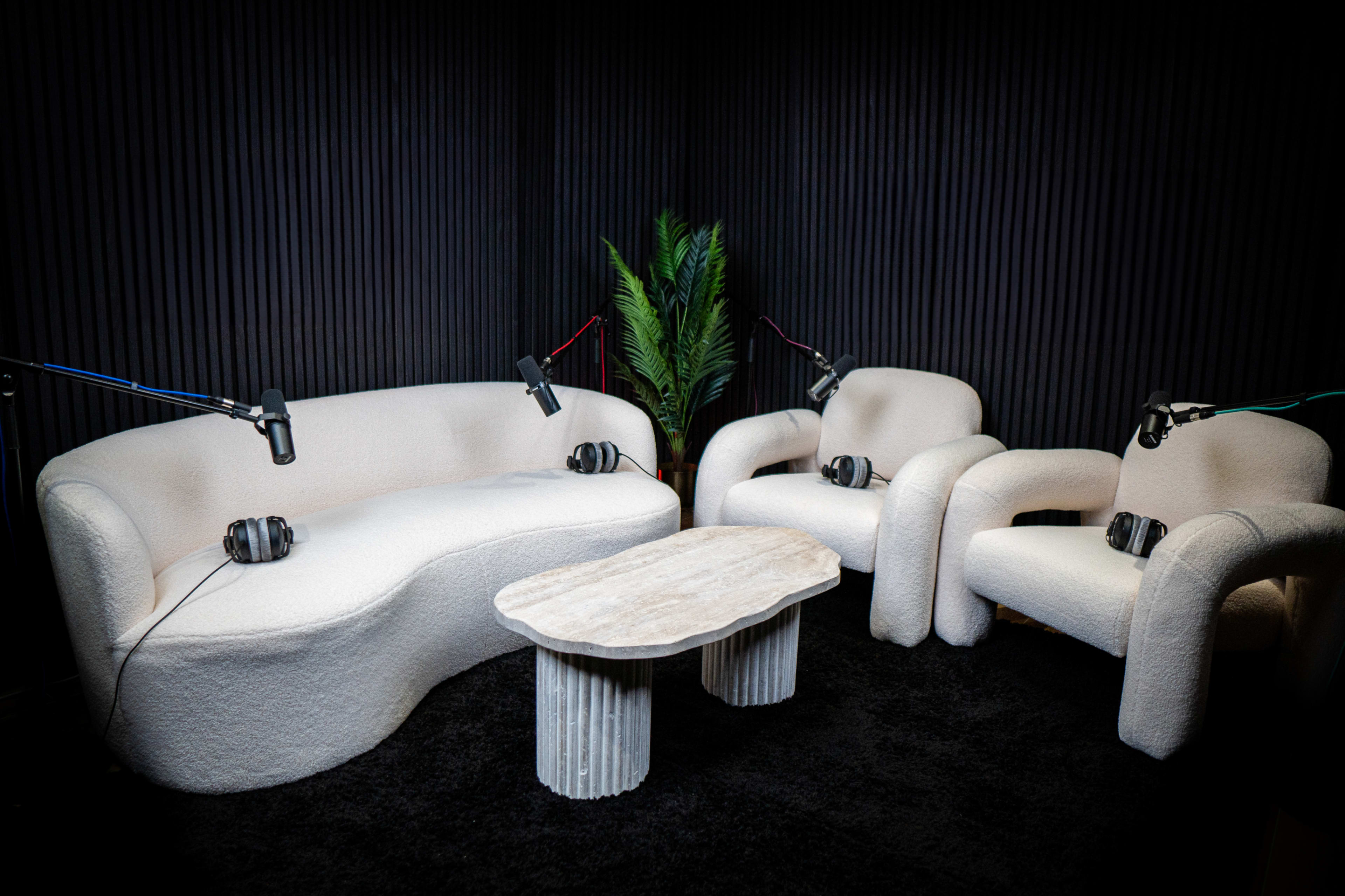 A cozy audio recording setup featuring a white, curved sofa and two matching armchairs, all with microphones and headphones positioned on them, alongside a stone coffee table.