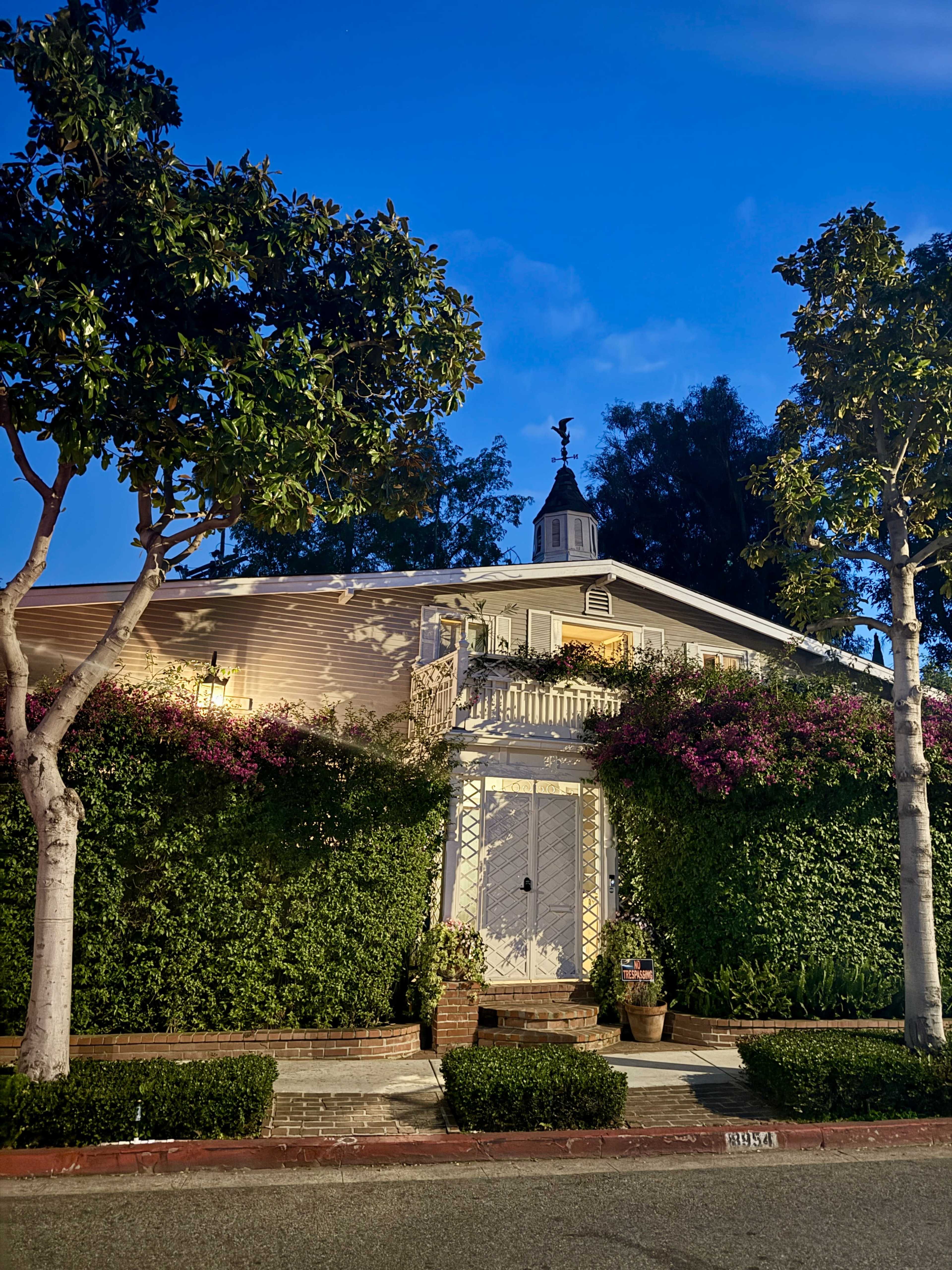 A two-story house with a cupola is surrounded by lush greenery and blooming flowers at dusk.