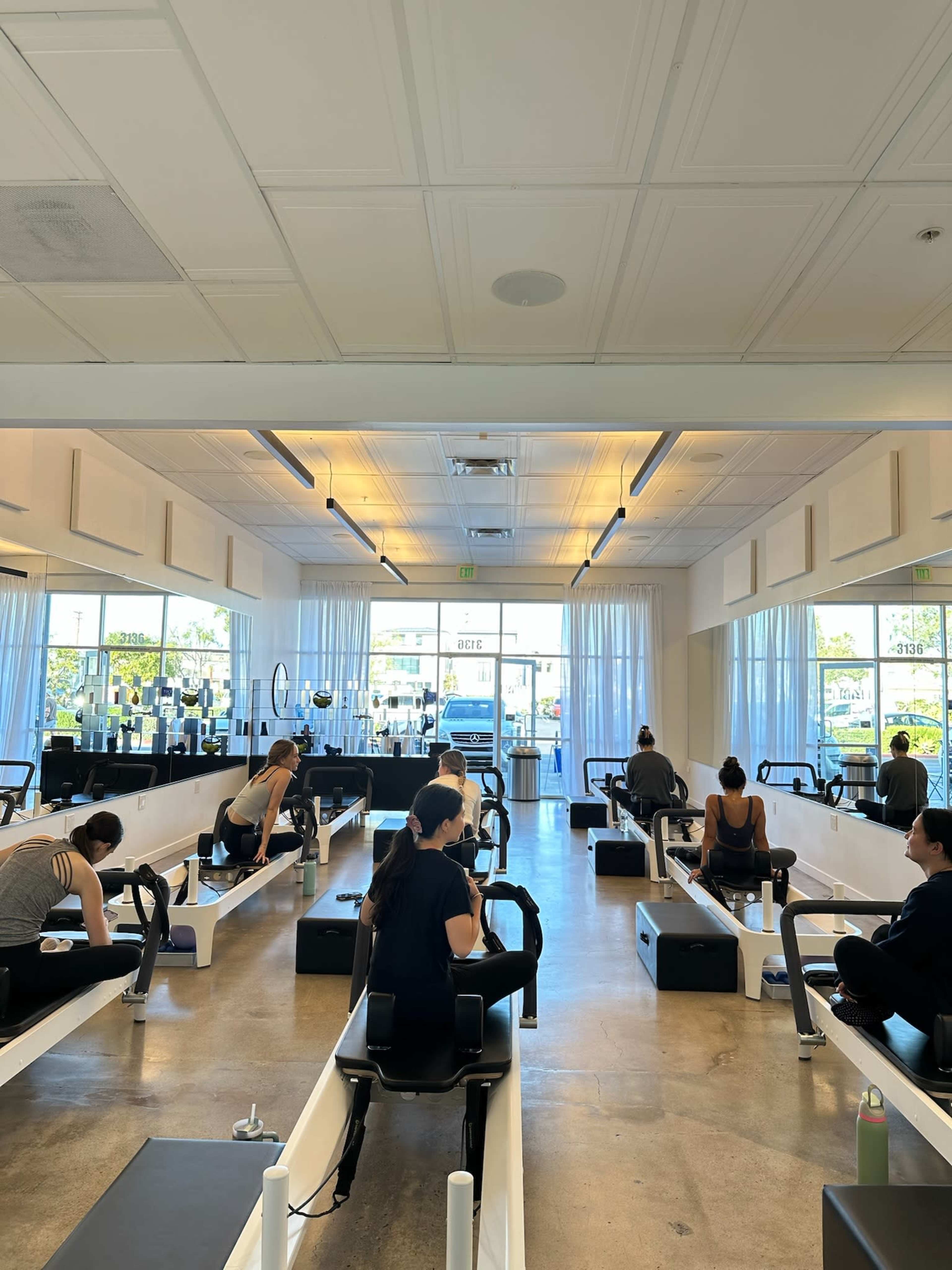 The image shows a fitness studio with several people participating in a workout on reformer machines, facing a large window with natural light streaming in.