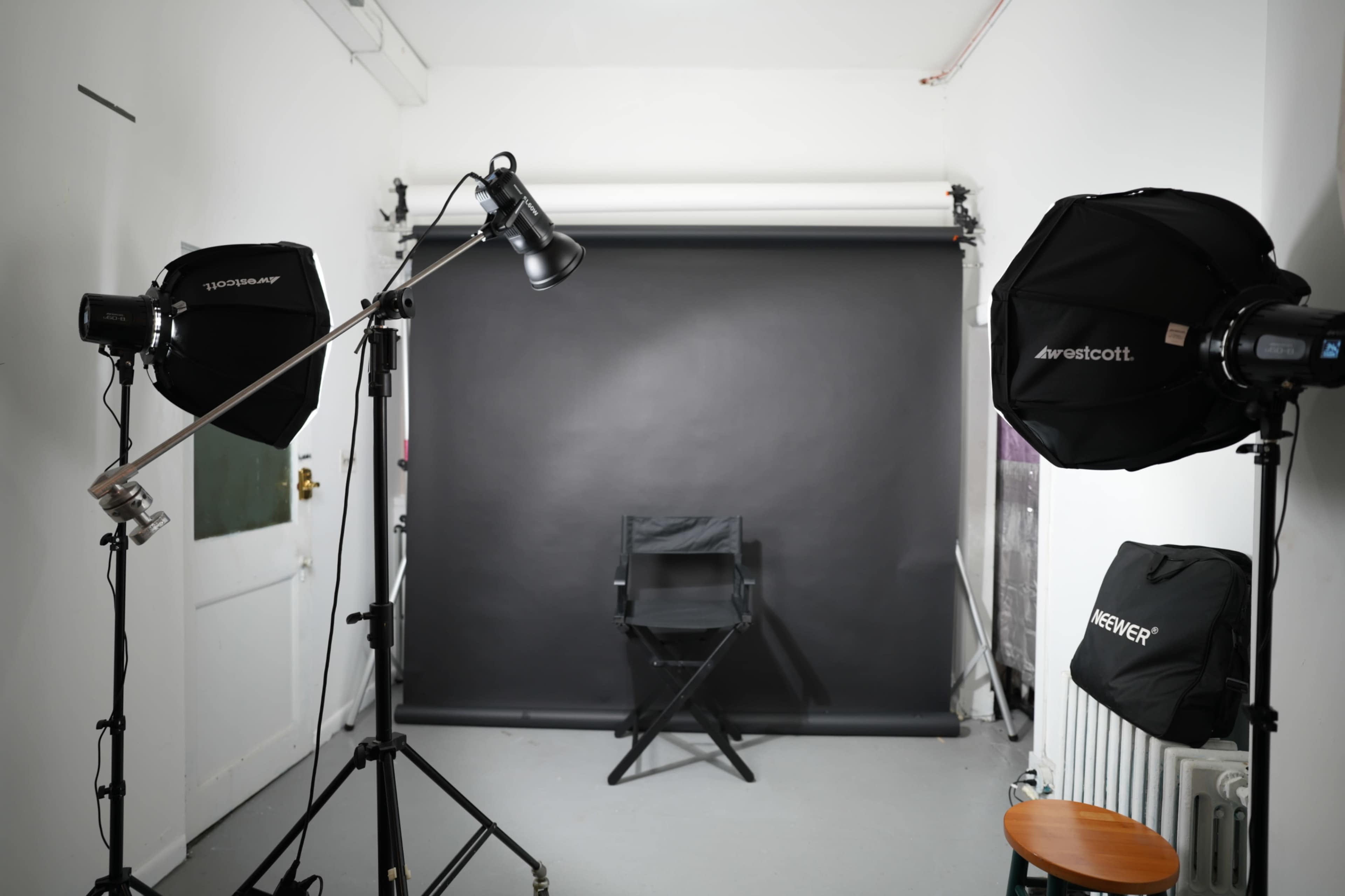 The image shows a photography studio setup with two softbox lights, a black backdrop, and a director's chair positioned in front of it.