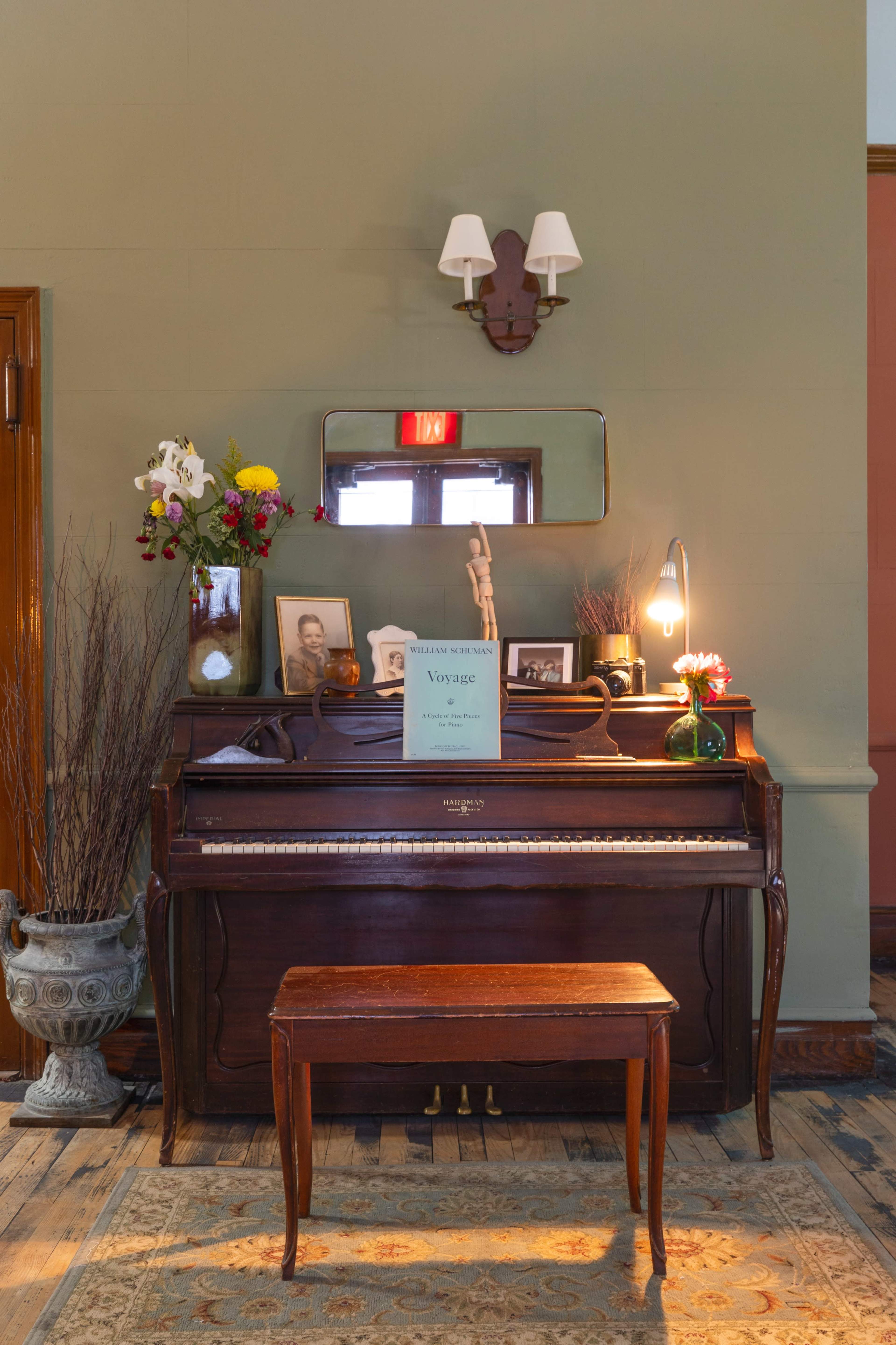 A wooden upright piano with a bouquet of flowers and a framed photo on top sits against a green wall, accompanied by a small table and a rug on the floor.