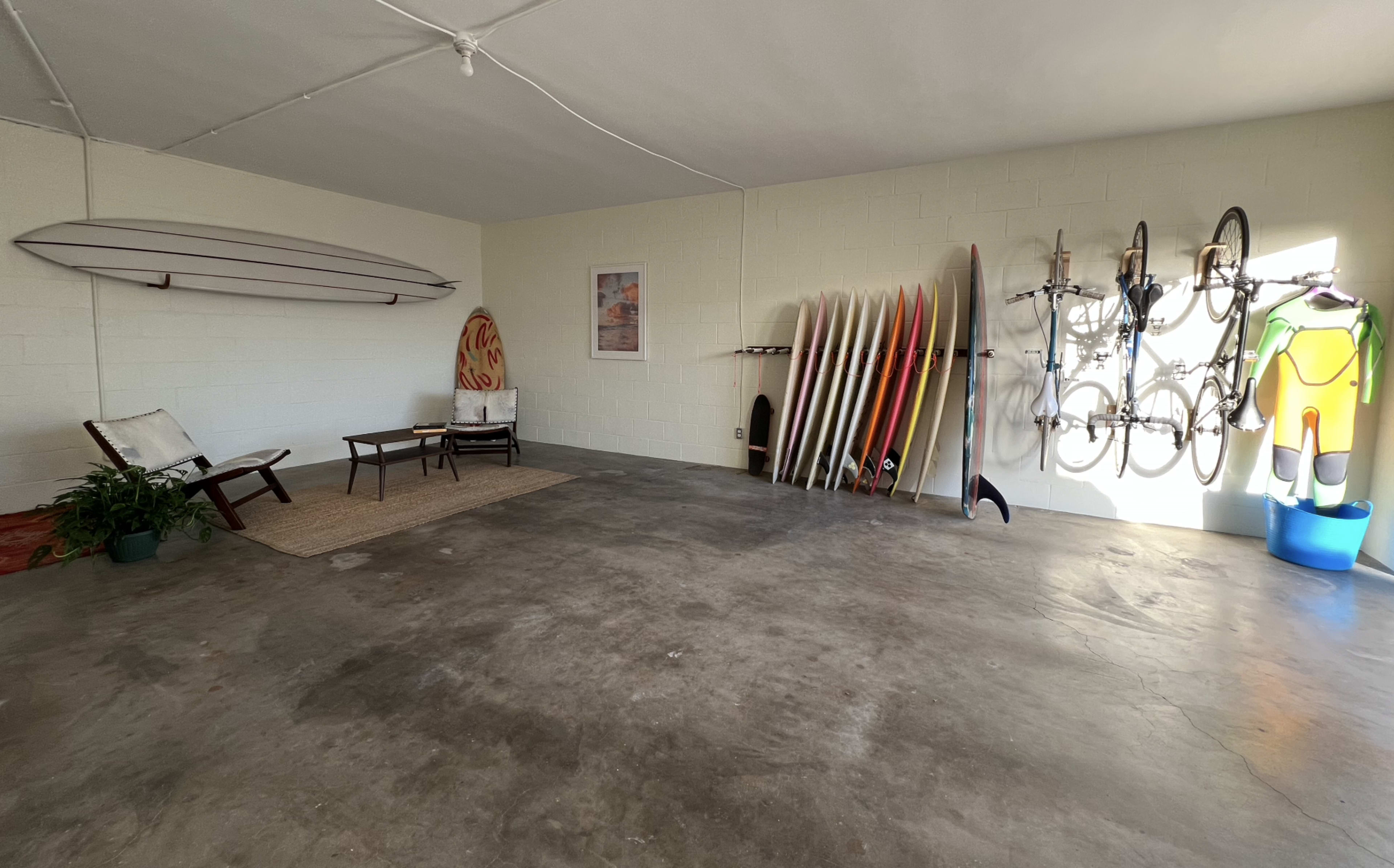 The image shows a spacious garage with surfboards and bicycles neatly arranged on the walls and a seating area consisting of a chair and small table.
