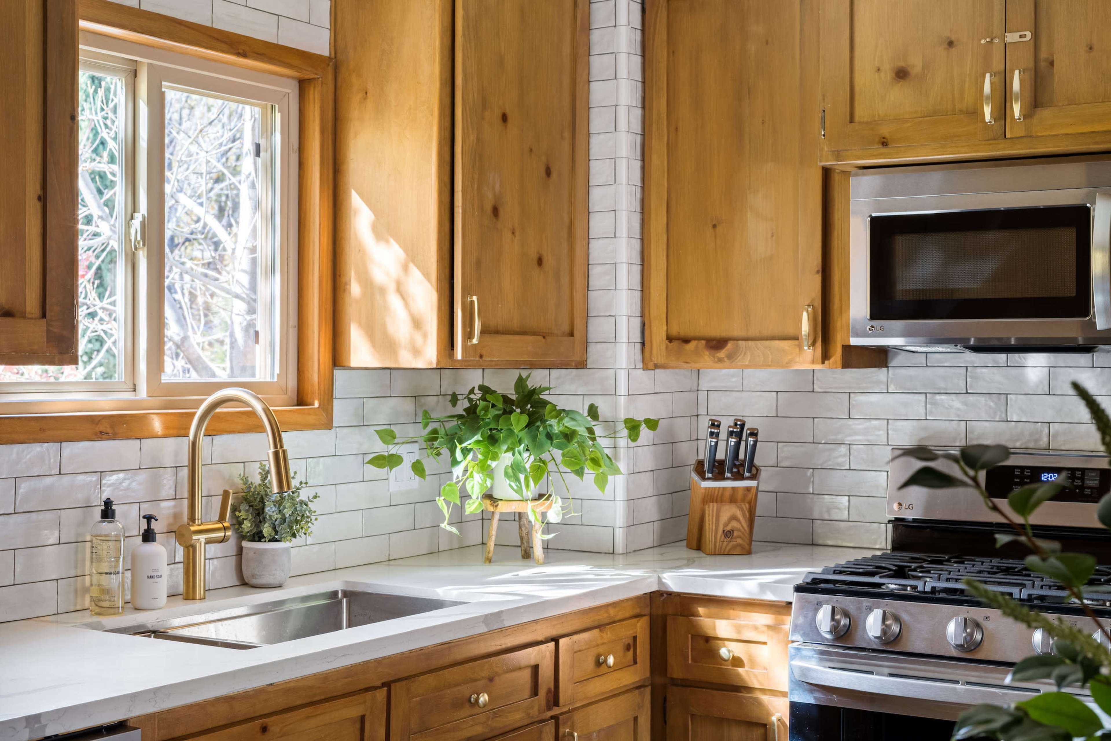 A well-lit kitchen features wooden cabinets, a stainless steel microwave, a gas stove, and a window overlooking greenery, with plants placed on the countertop.