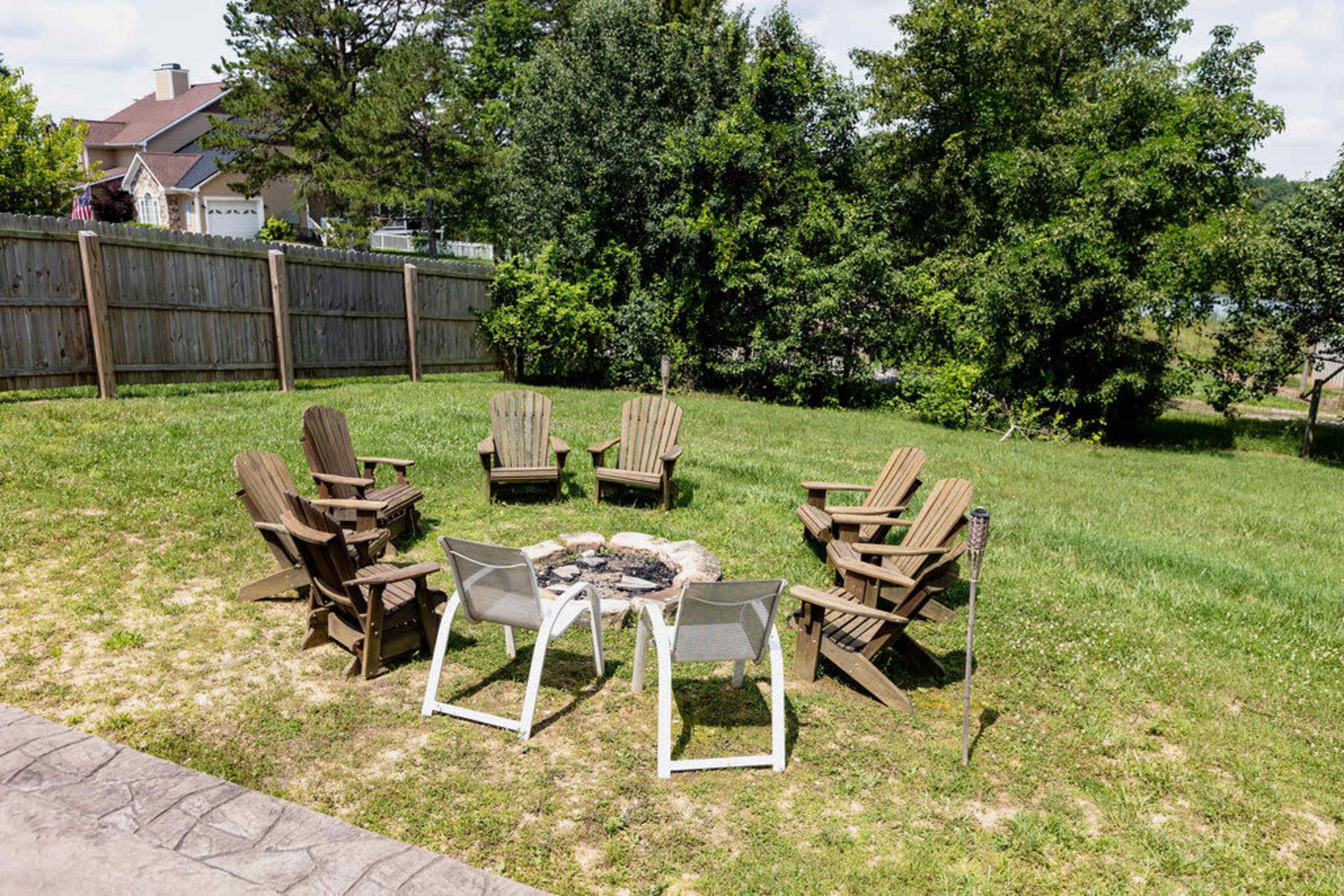 A circular arrangement of wooden chairs surrounds a fire pit in a grassy yard, enclosed by a wooden fence and trees.