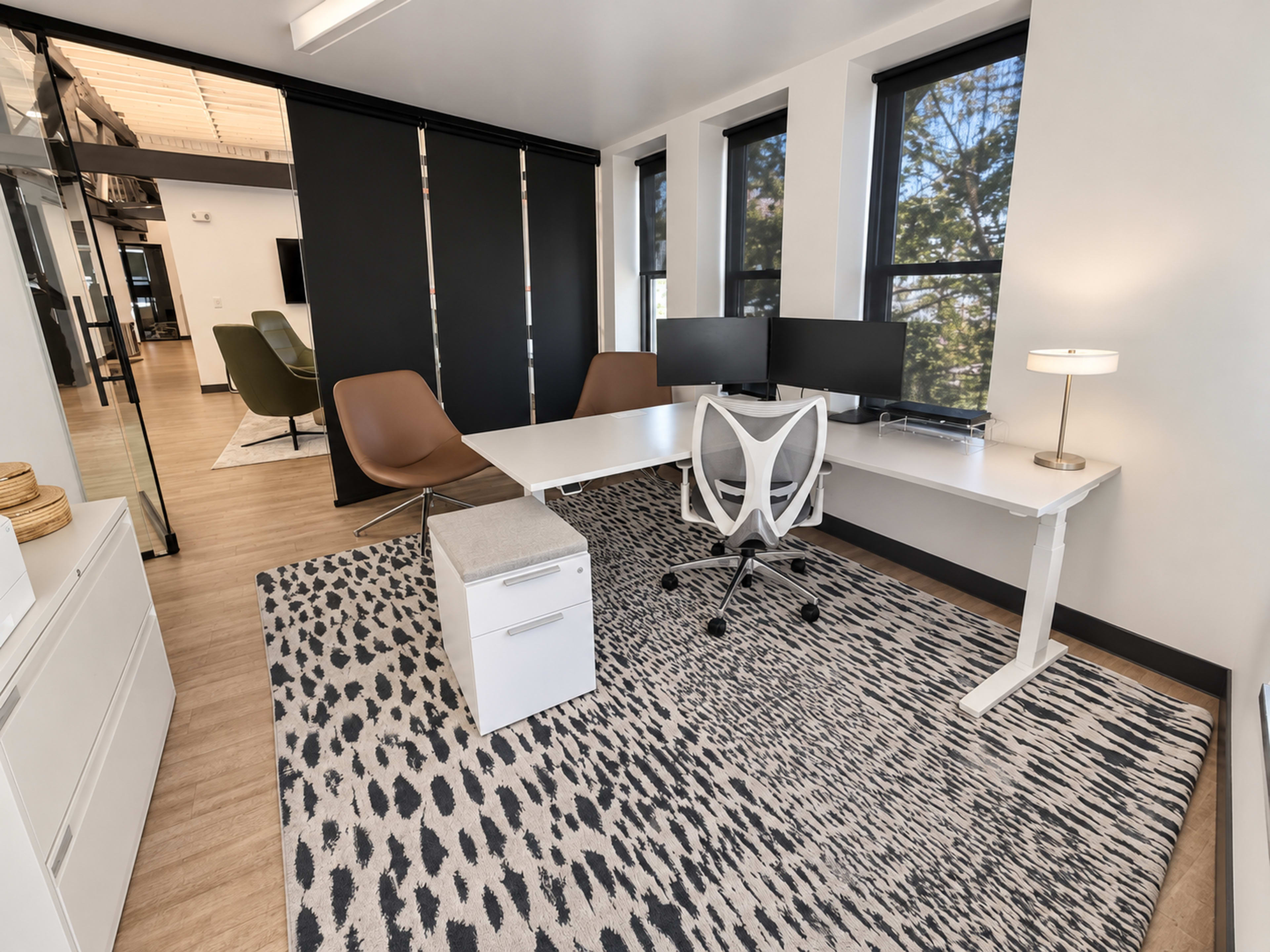 The image shows a modern office space featuring a white desk with dual monitors, a swivel chair, a light fixture, a small storage unit, and large windows allowing natural light to enter.
