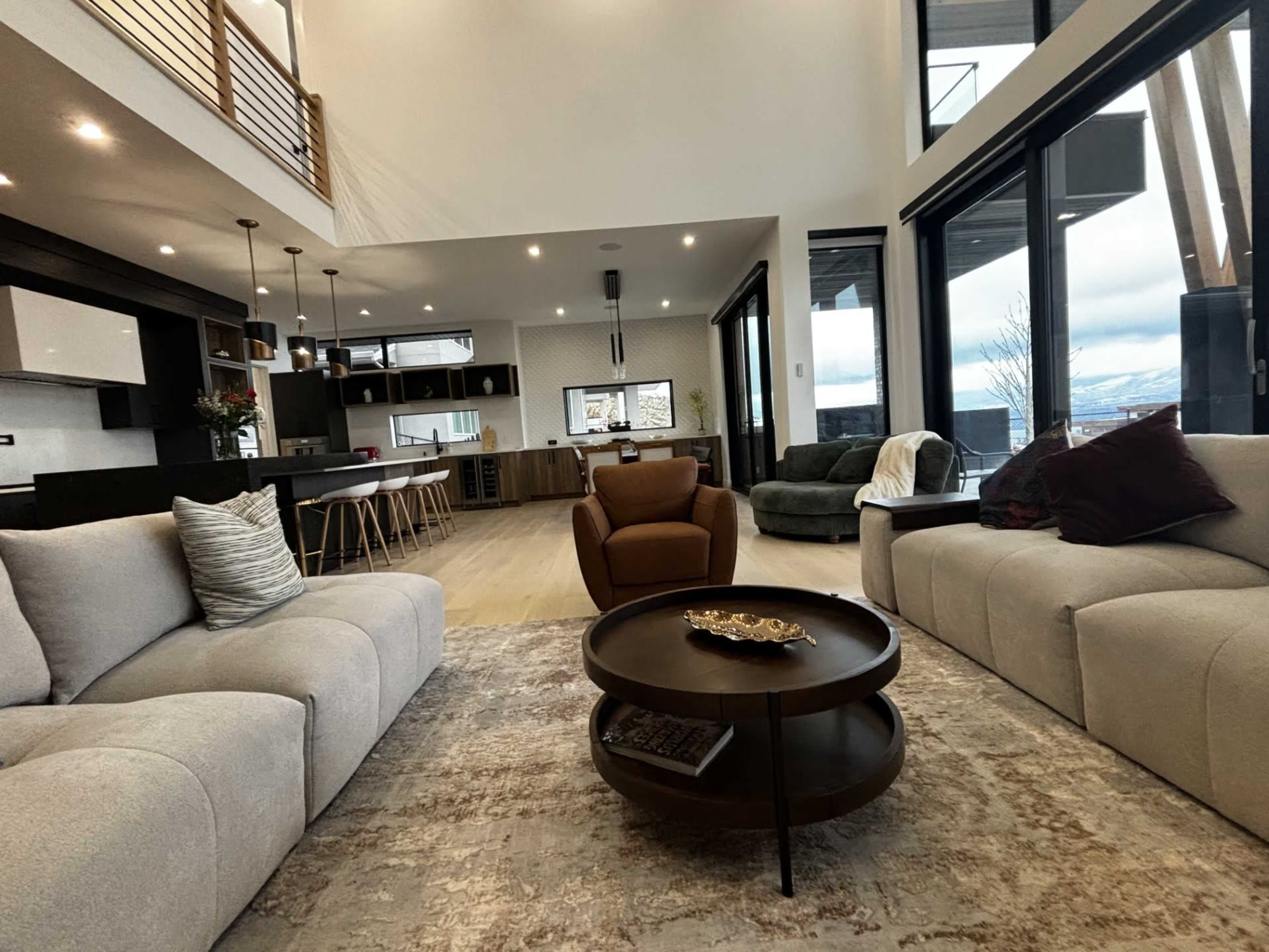 The image shows a modern living room with two sofas facing a coffee table, a brown armchair, and large windows overlooking a scenic view.