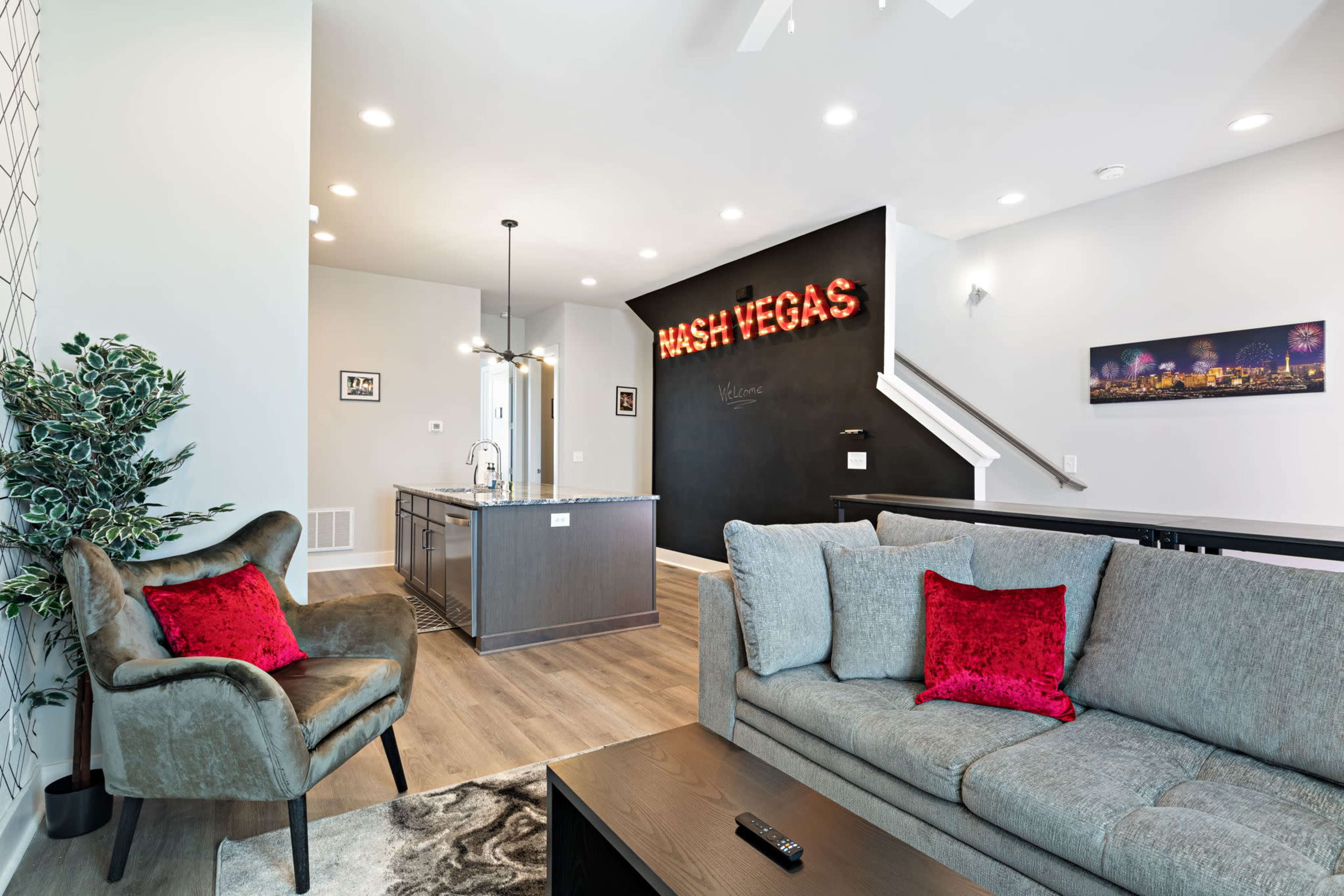 The image shows a modern living room with a gray sofa and an armchair, accented by red pillows, next to a kitchen island with a blackboard wall displaying "Welcome to Las Vegas."