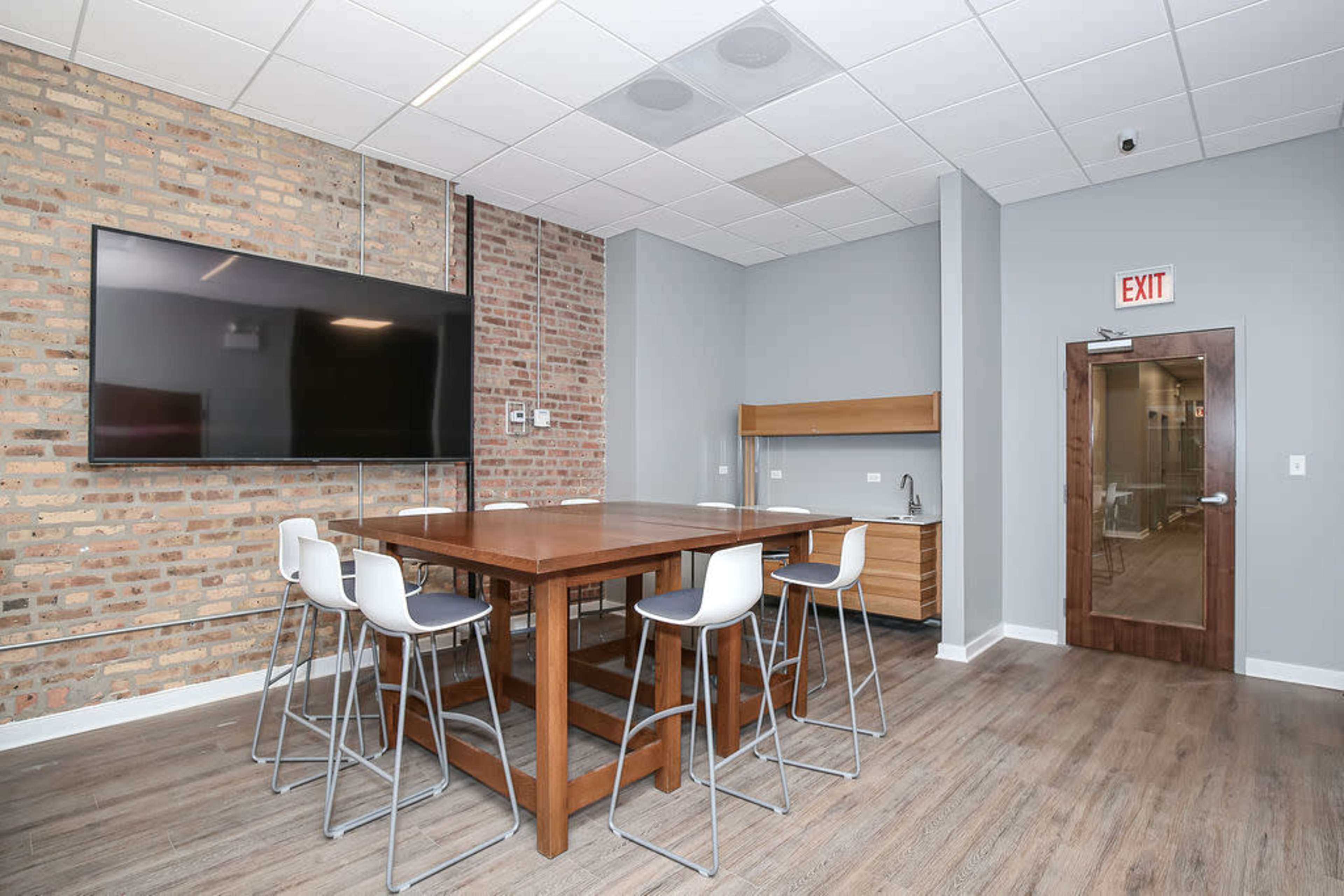 A modern meeting room with a wooden table, high-backed chairs, a large wall-mounted screen, and a small kitchenette area.