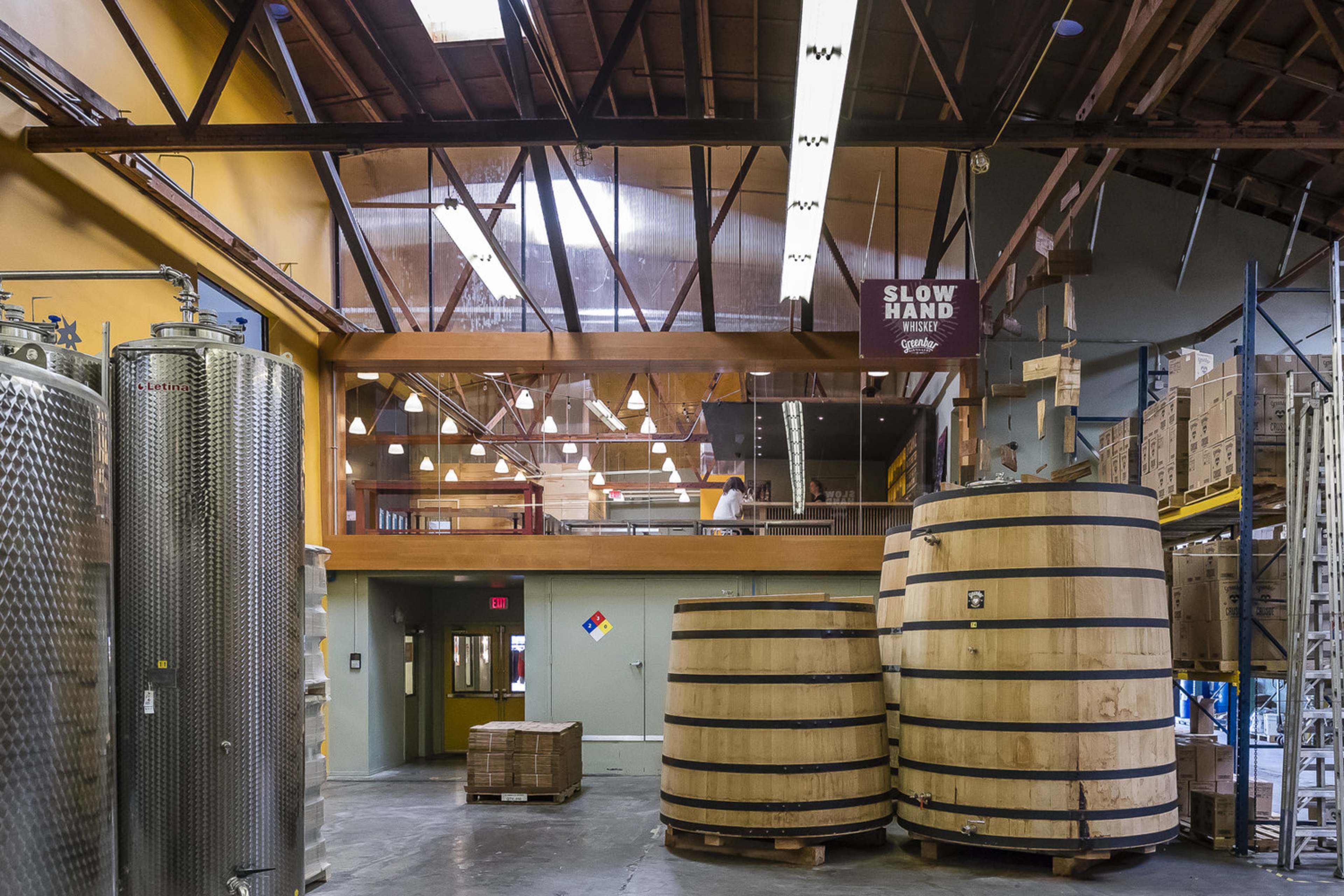 A modern industrial space with large wooden fermentation barrels, stainless steel tanks, and a mezzanine level overlooking the area.