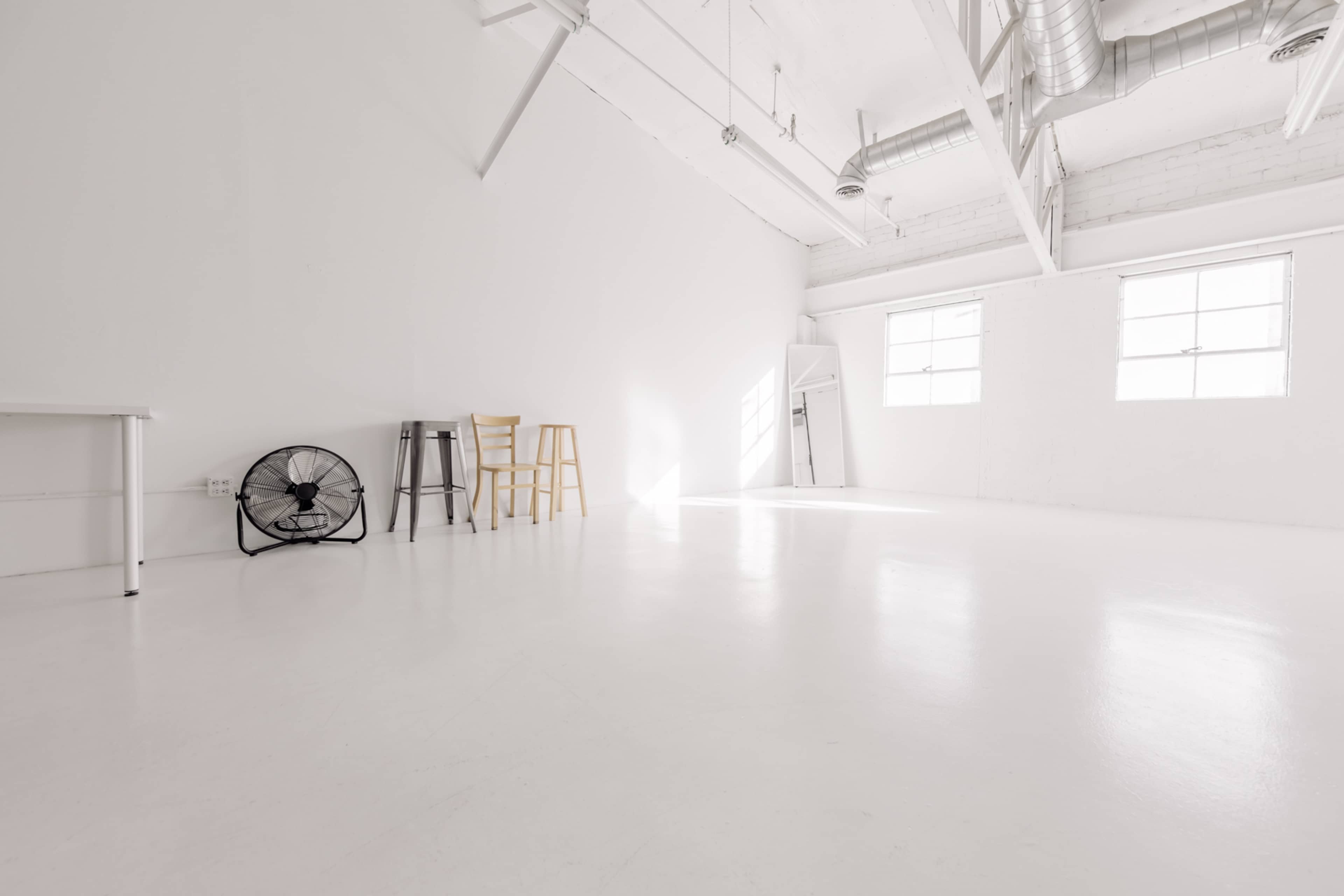 A spacious, empty room with white walls and a polished floor, featuring a fan, a table, and two tall chairs.