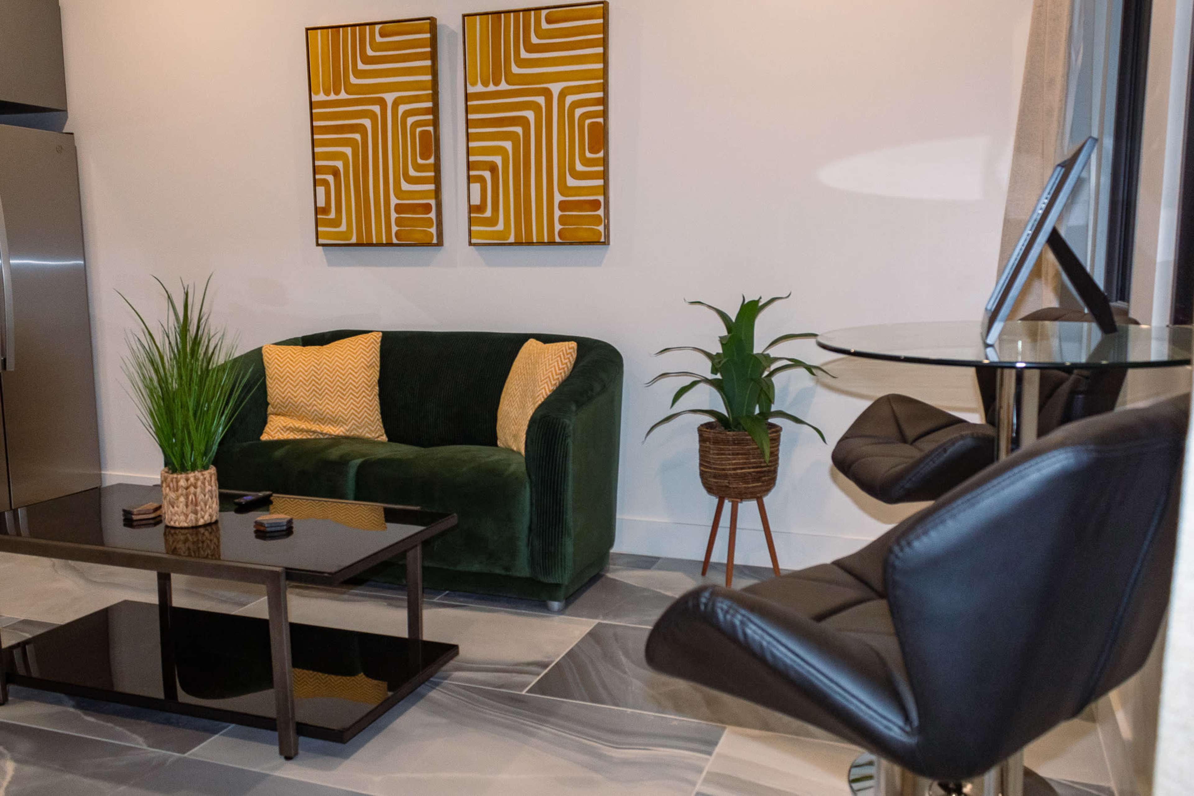 The image shows a modern living area featuring a green sofa, two abstract art pieces on the wall, a glass-topped coffee table, and a small round dining table with black chairs.