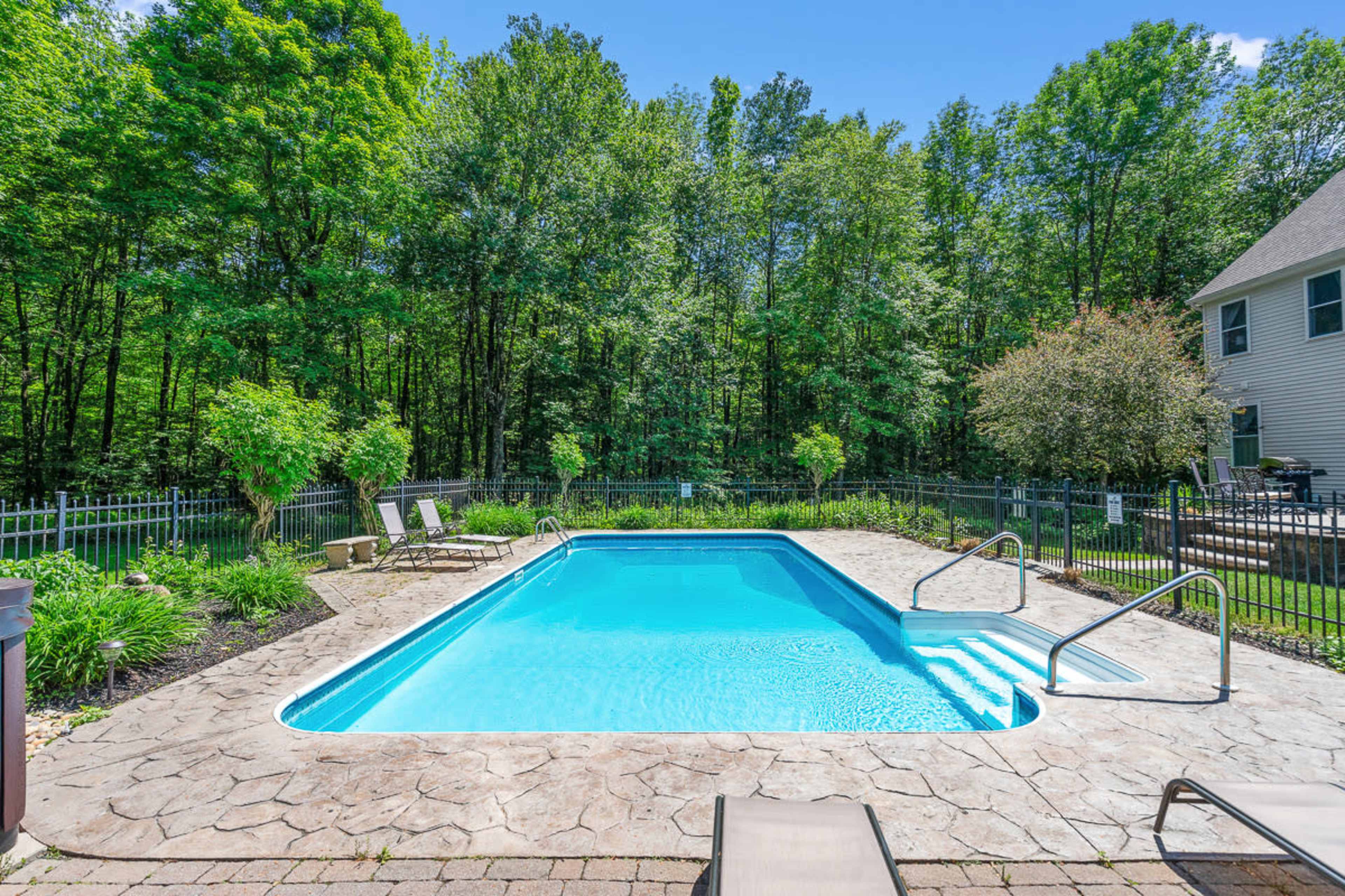 A rectangular swimming pool with a stone deck is surrounded by a fenced, green backyard and trees.