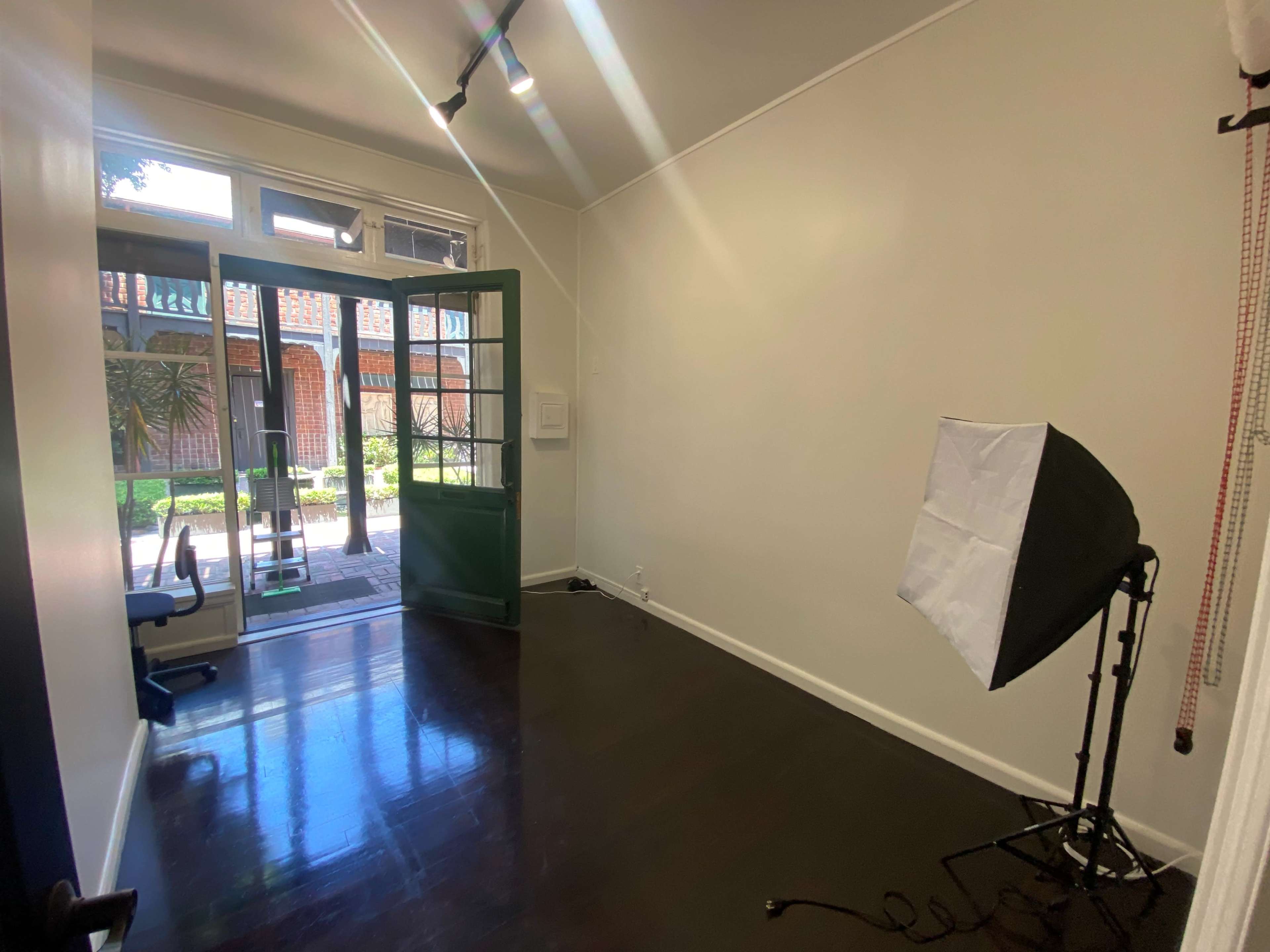 The image shows a spacious, empty room with a green double door leading to an outdoor space, featuring a lighting setup in one corner.