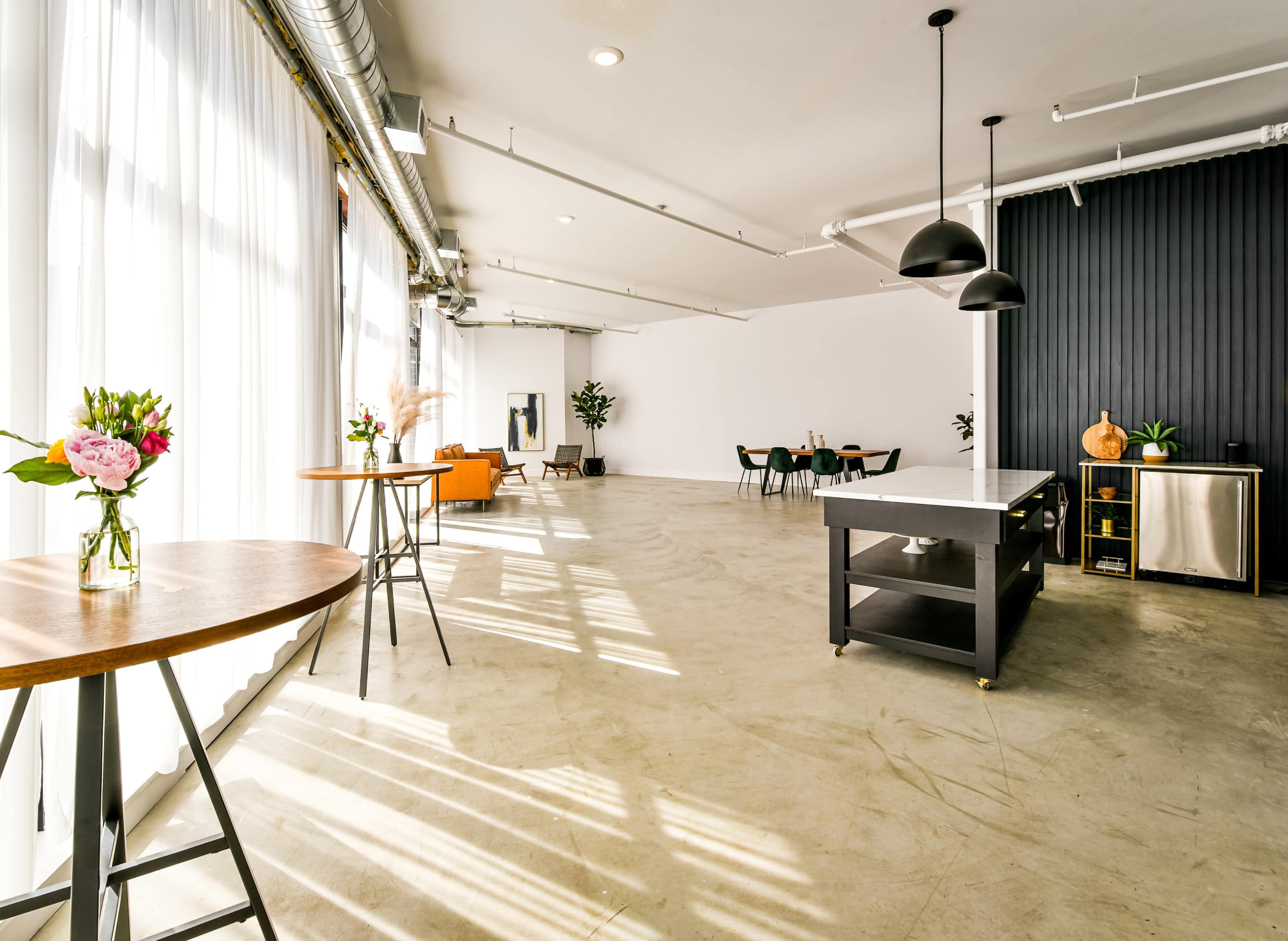 A spacious, minimalist interior with large windows, natural light flooding in, and a mix of wooden tables, green chairs, and a dark feature wall.