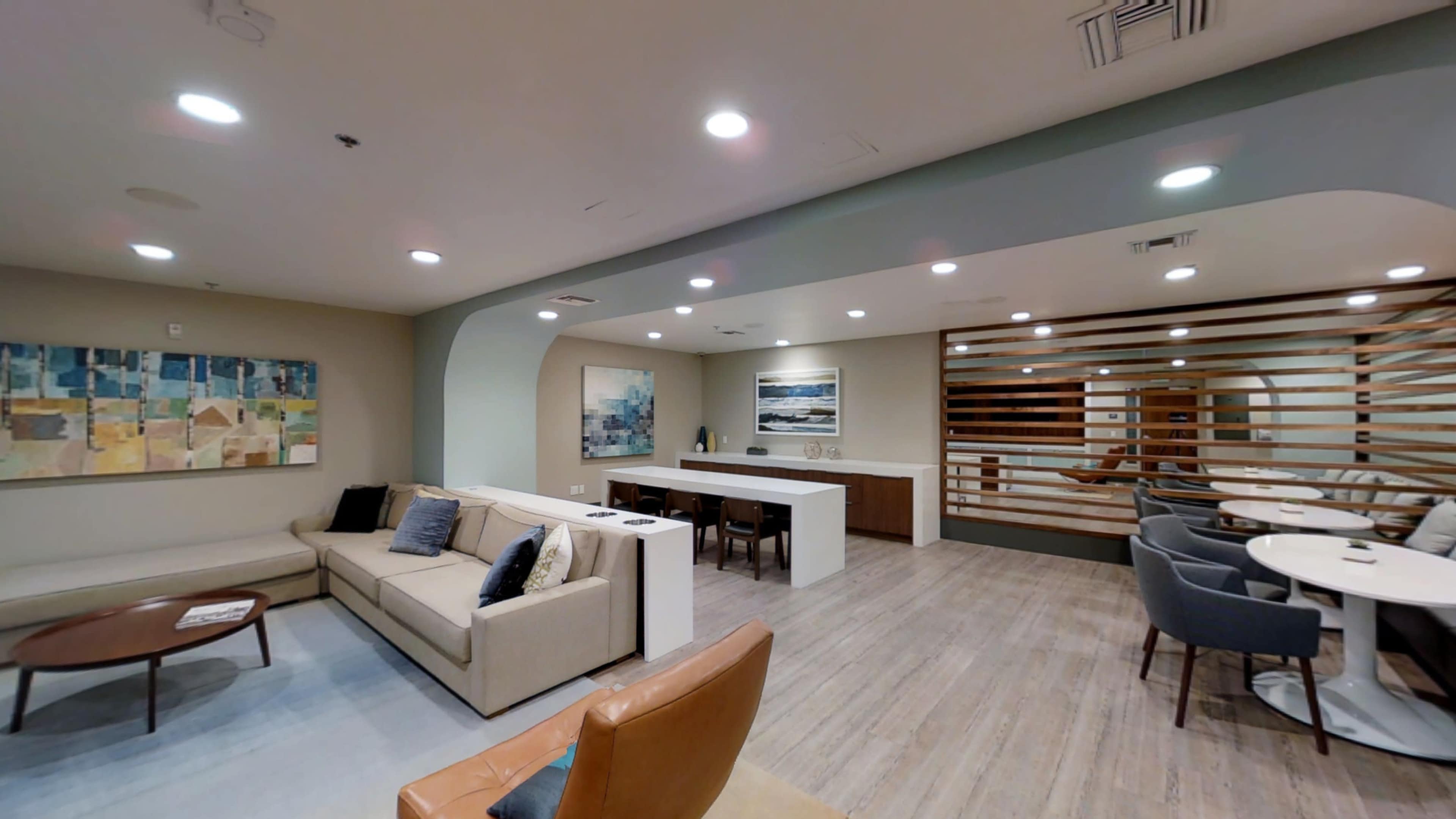 A modern lounge area featuring a beige sofa, a wooden coffee table, and a dining space with light-colored furnishings and artwork on the walls.