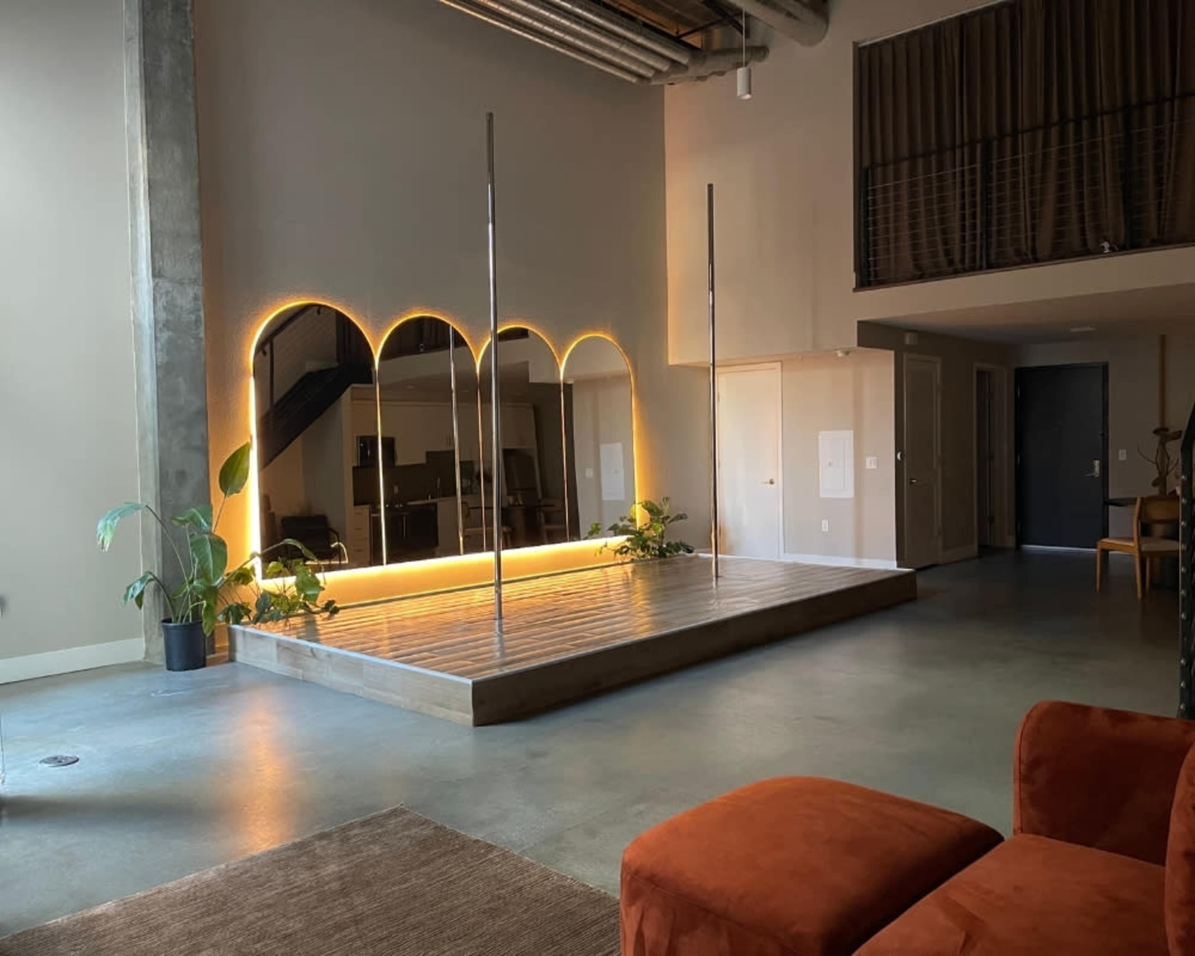 The image shows a spacious interior featuring a wooden platform with two vertical poles, a large mirror, and modern furnishings, illuminated by soft lighting.
