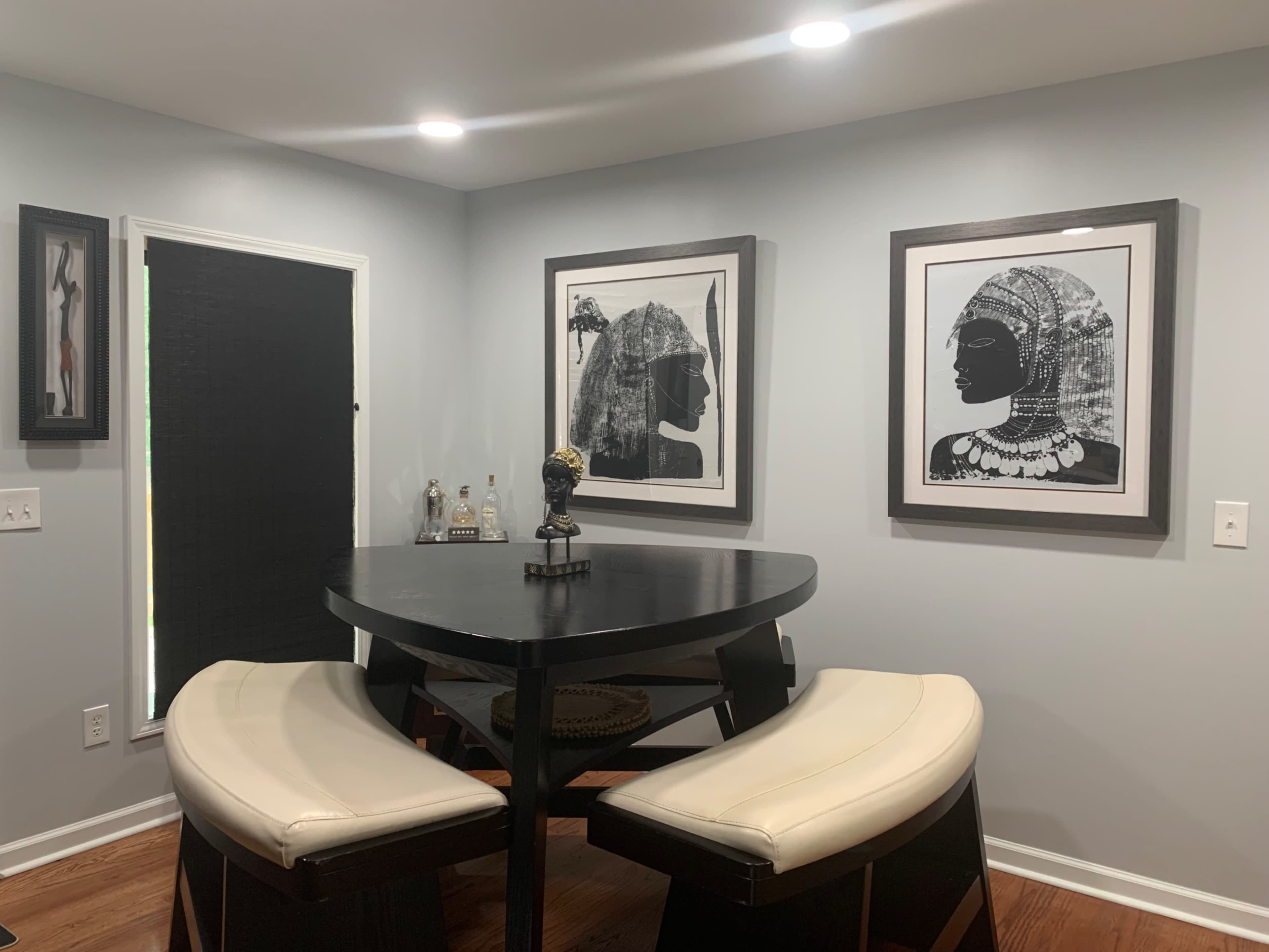 A round black table with two white cushioned chairs is positioned next to a wall featuring two framed black-and-white artworks of profiles.