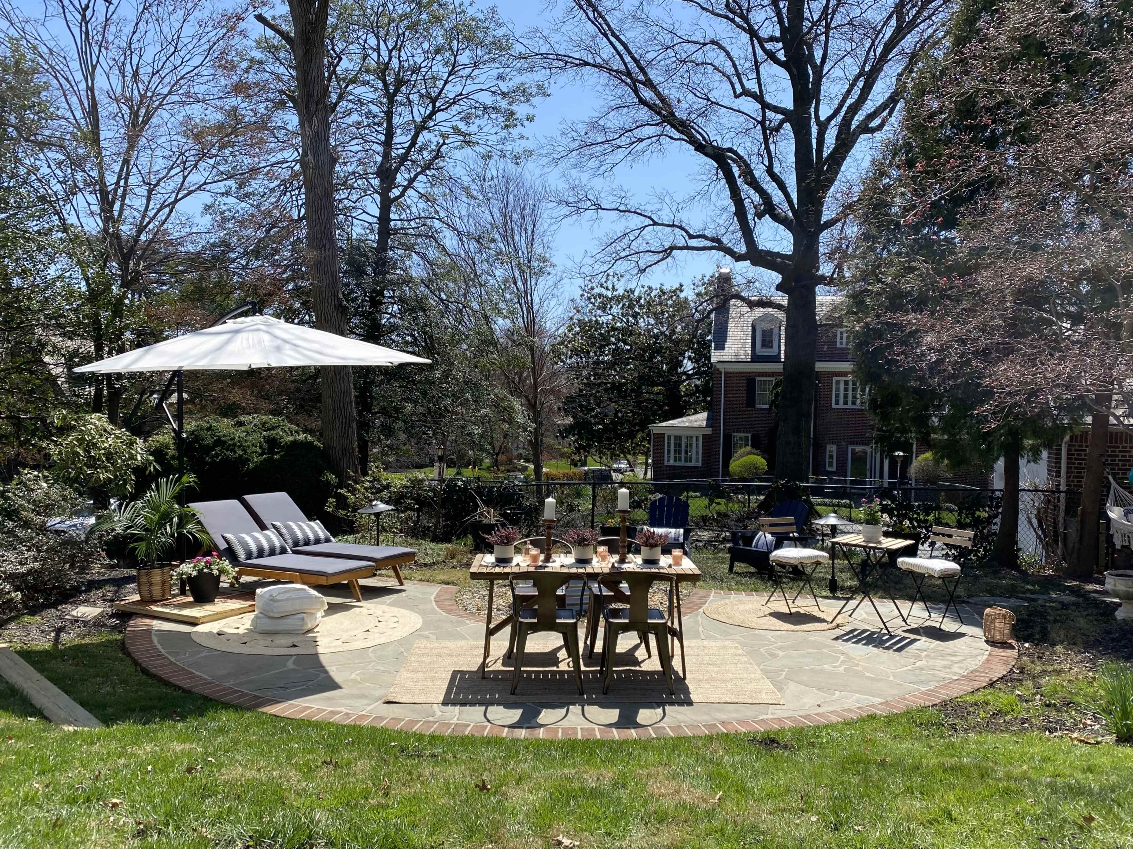 A spacious outdoor patio features a dining table set for several guests, lounge chairs, an umbrella, and greenery surrounding a stone circular area.