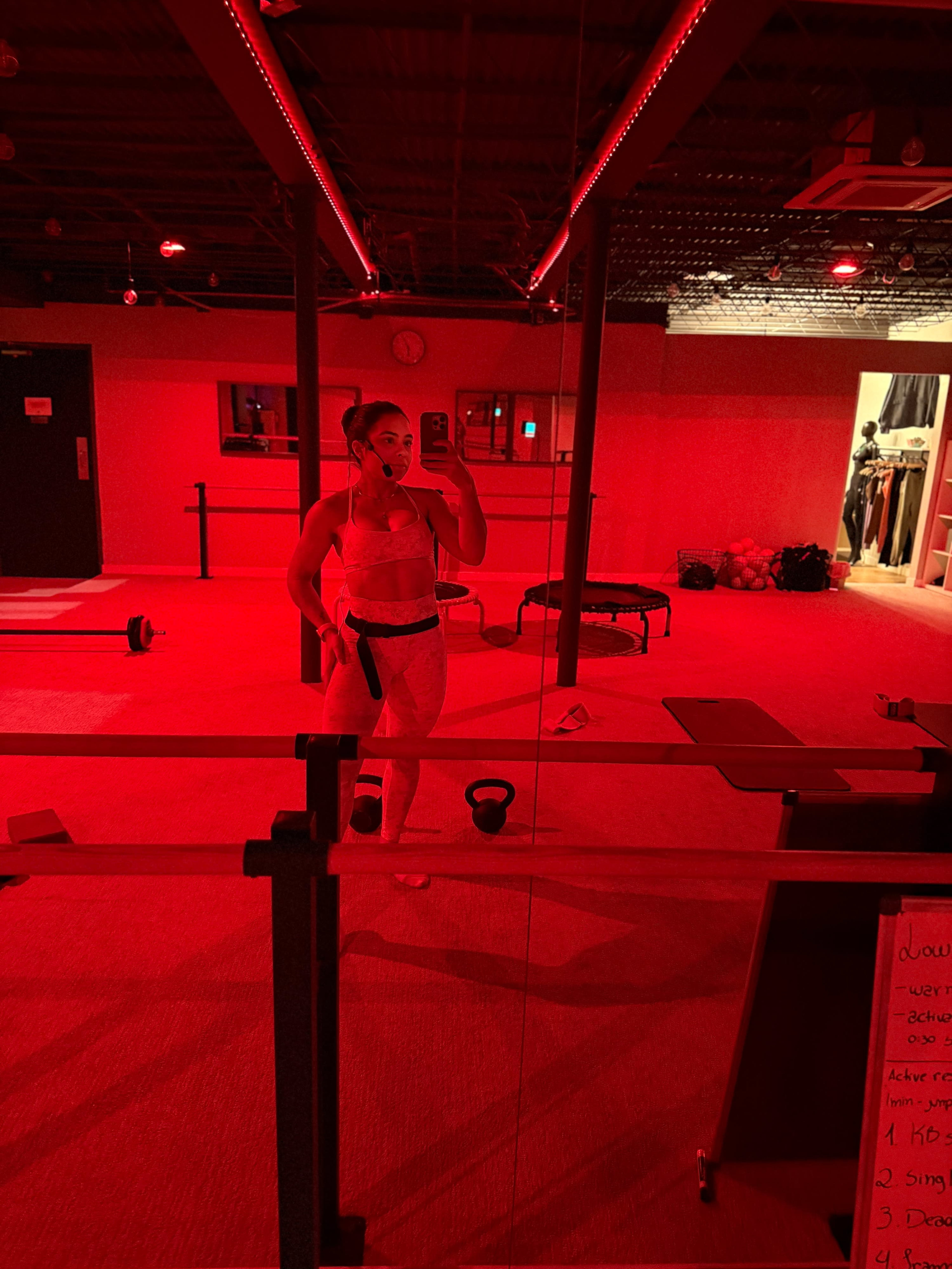 A person is standing in a gym with red lighting, taking a selfie while surrounded by exercise equipment.