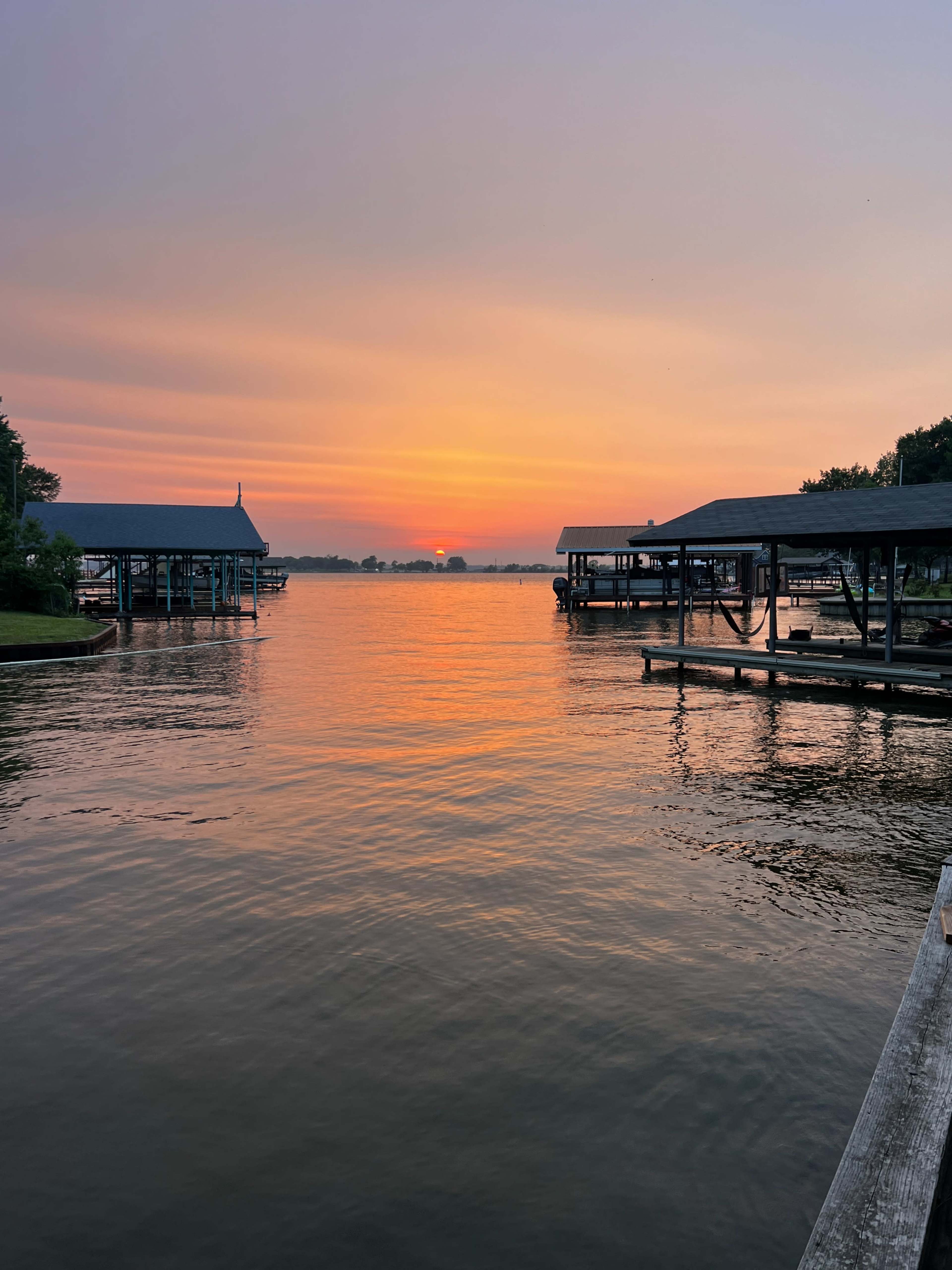 A calm lake at sunset, with silhouettes of boat docks on either side and a sun setting in the distance.