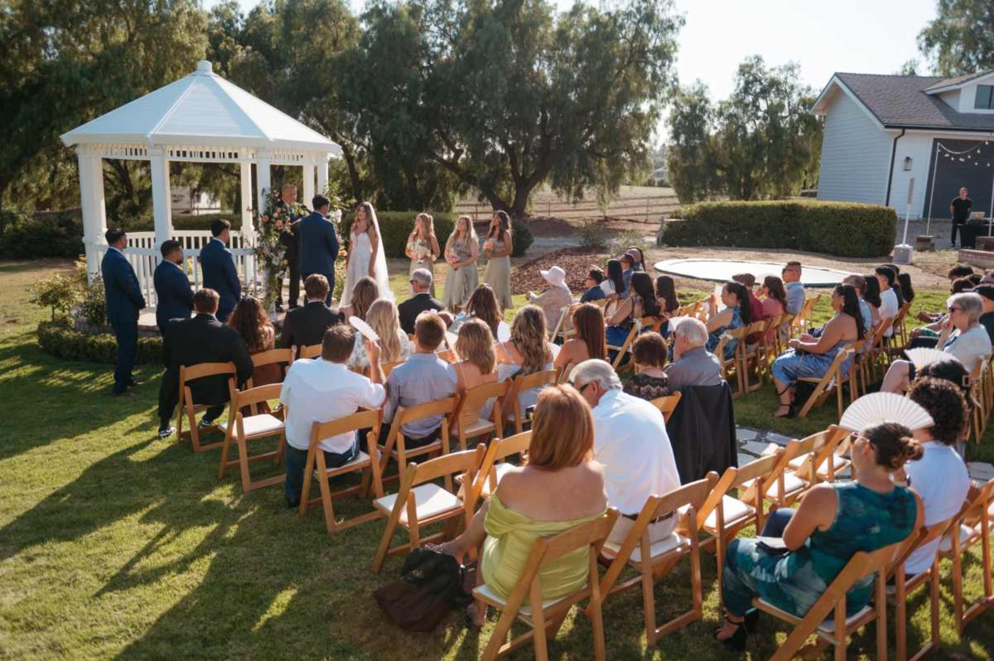 A wedding ceremony takes place outdoors under a gazebo, surrounded by guests seated on wooden chairs.