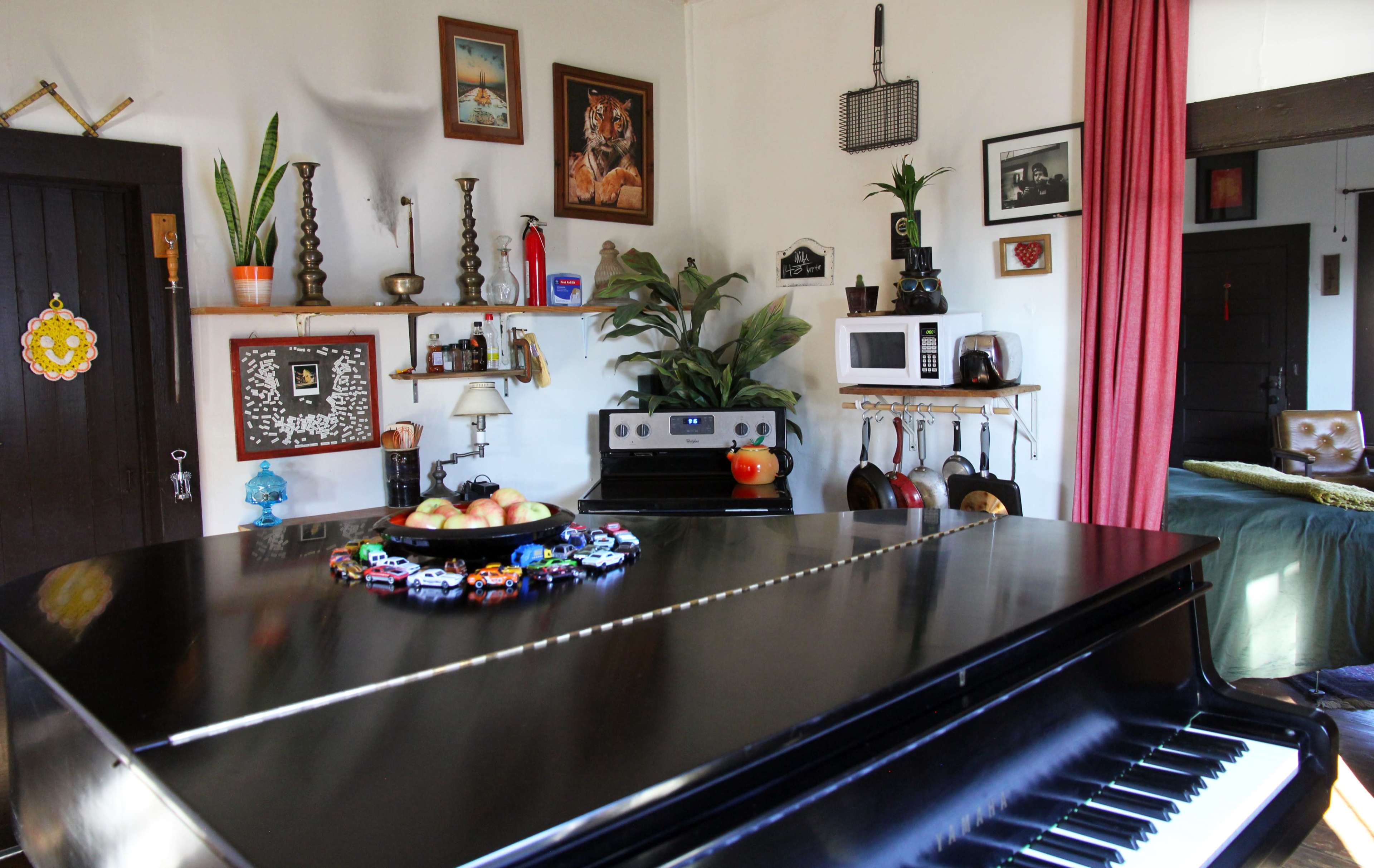 A black grand piano is positioned in the foreground of a cozy kitchen and living area, decorated with various plants, artwork, and kitchen utensils.