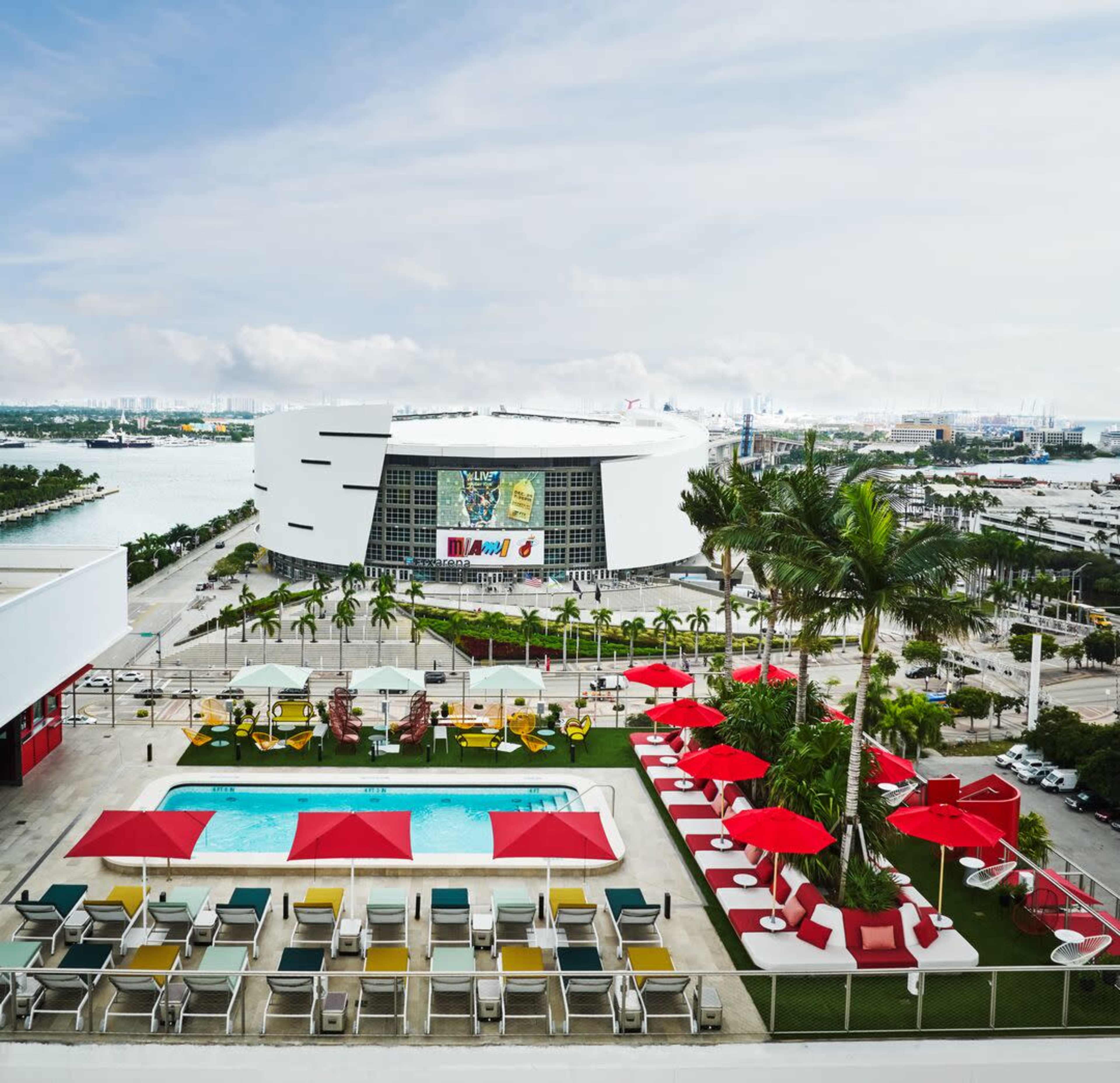 A rooftop pool area with red umbrellas and lounge chairs overlooking a sports arena and a waterfront in a cityscape.