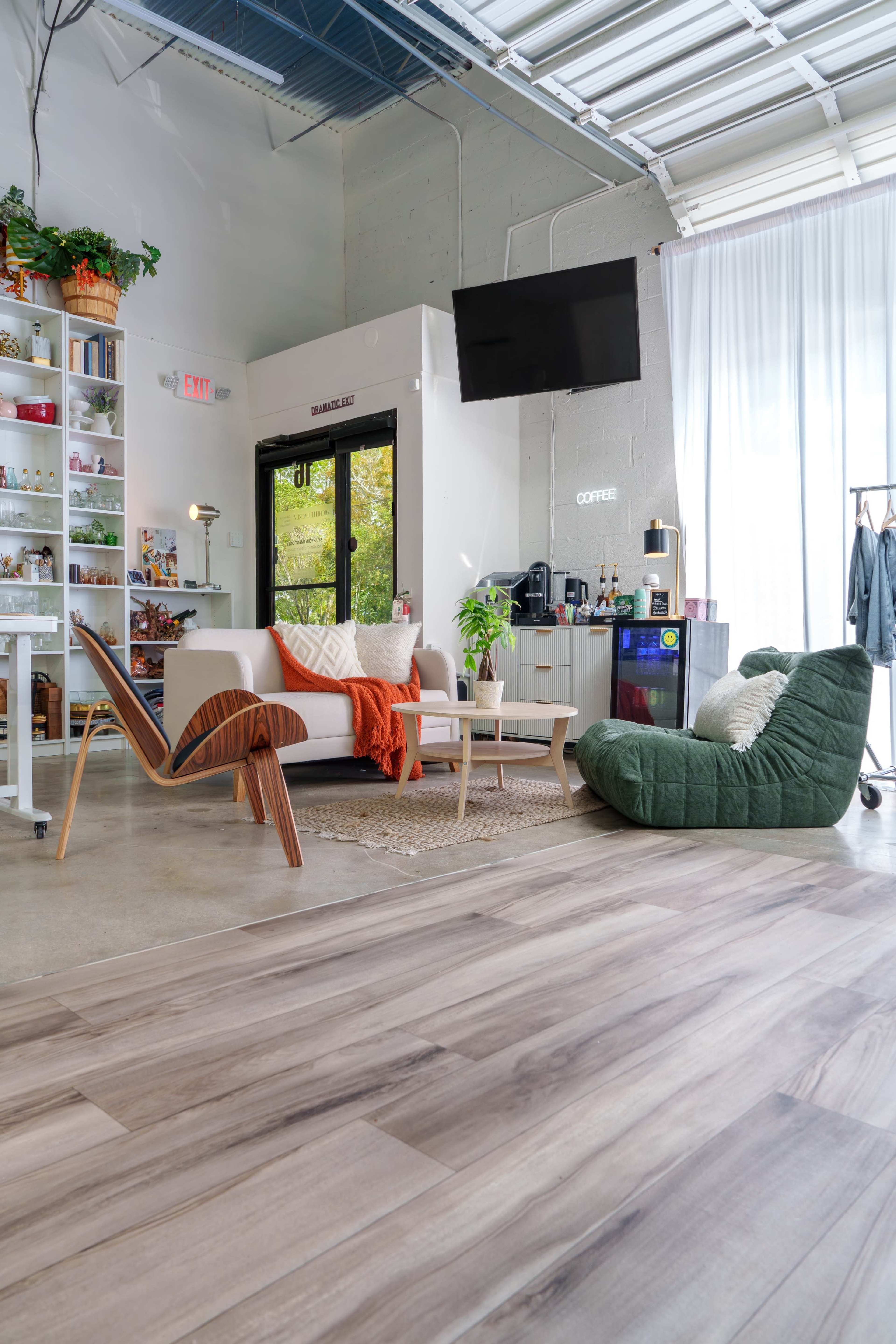 The image shows a modern interior space featuring a cozy seating area with a white sofa, a green bean bag chair, a coffee station, and shelves filled with various items.
