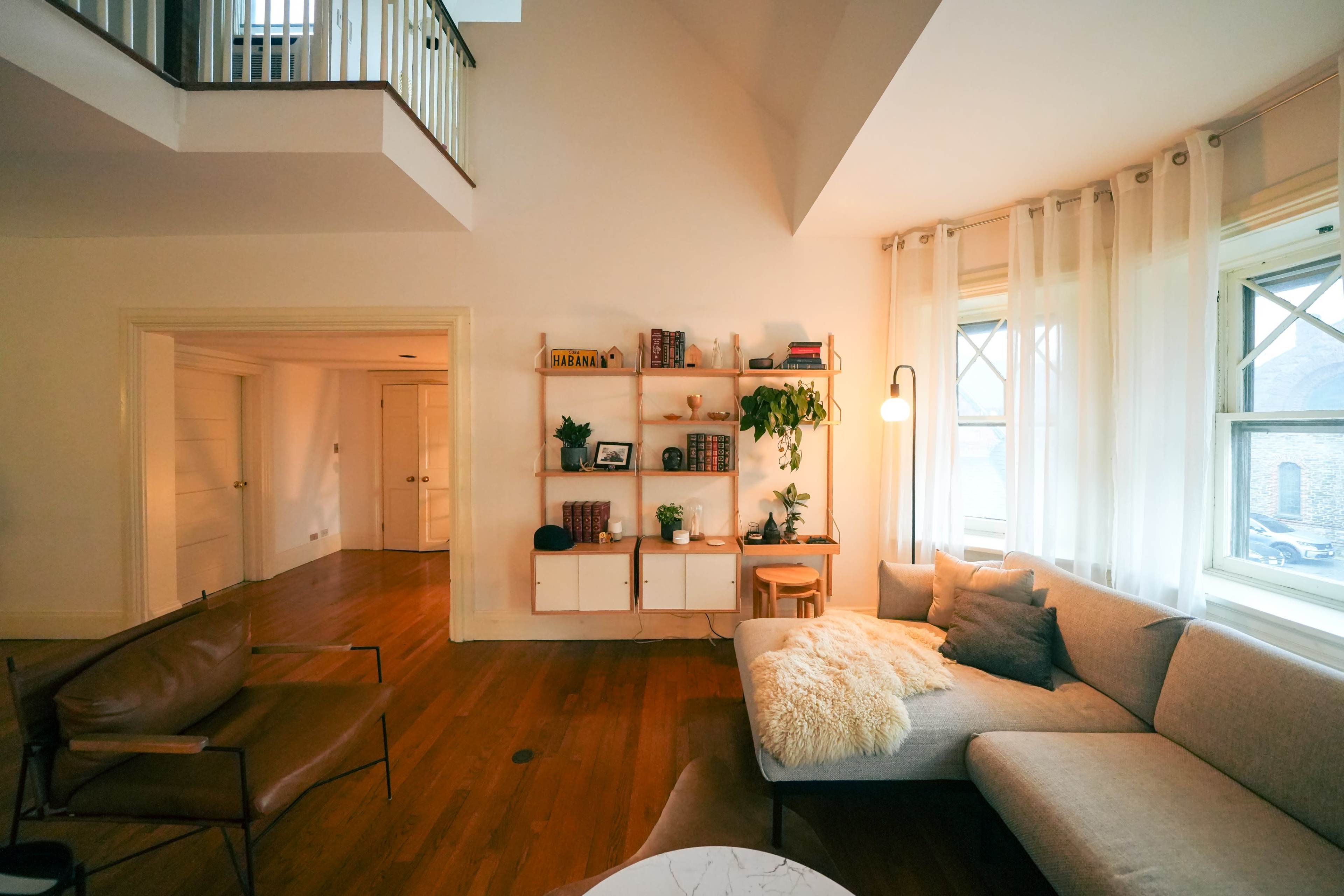A modern living room with a sectional sofa, a bookshelf filled with plants and books, and large windows letting in natural light.