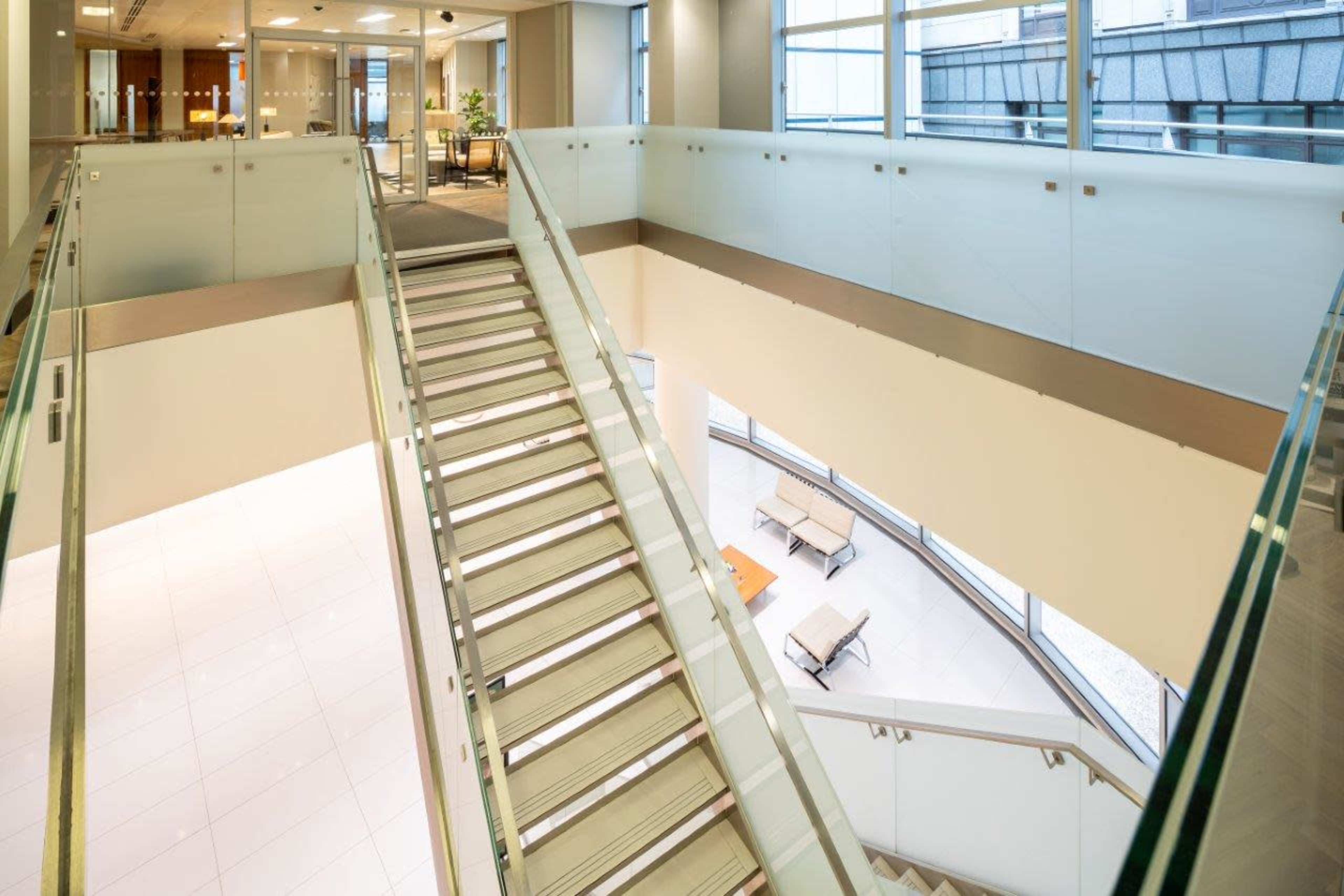 The image shows a modern interior with a glass staircase leading to an open area that includes seating and large windows.