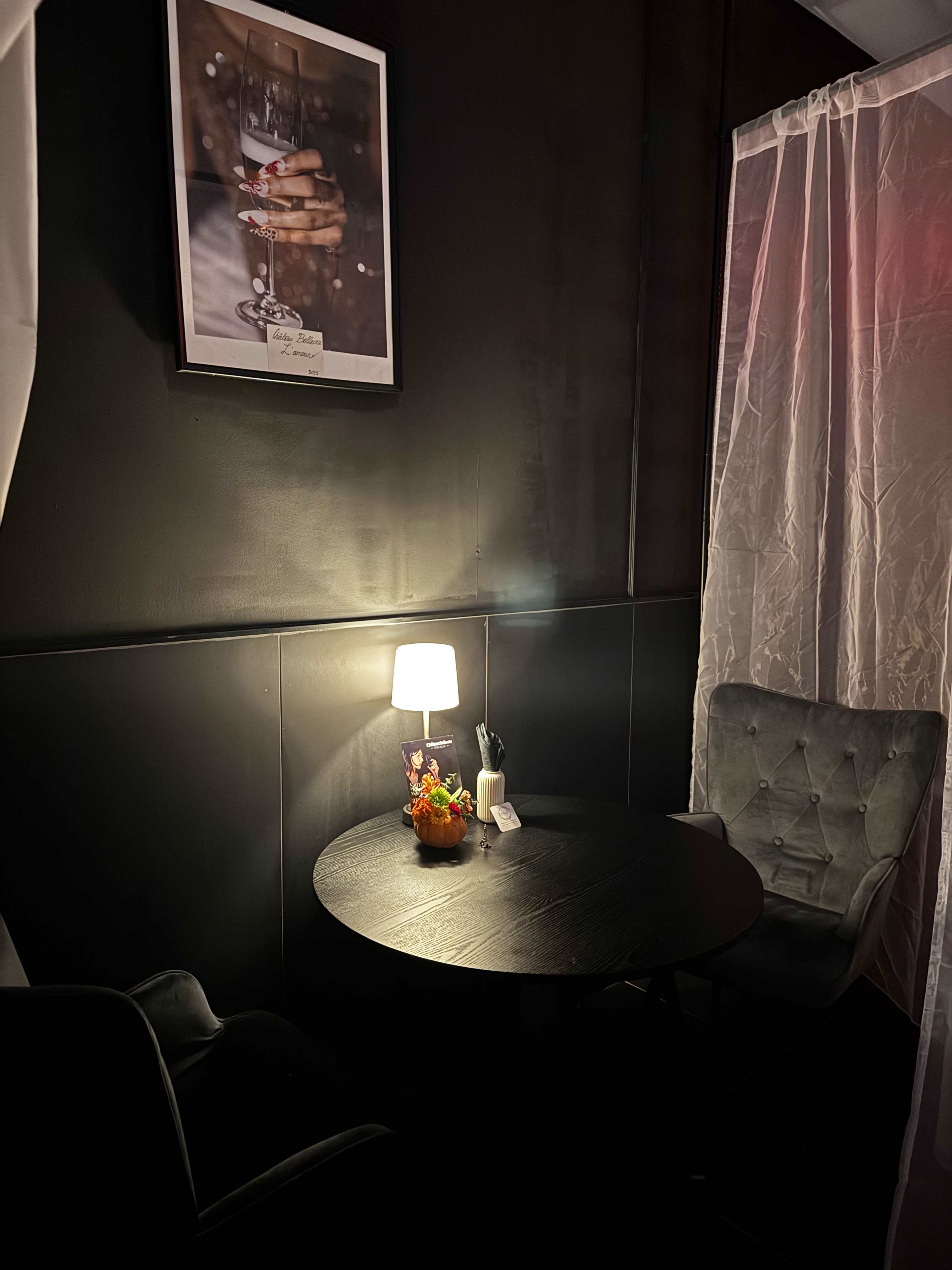 A small, dimly lit corner with a round table, two chairs, a table lamp, and a fruit arrangement, surrounded by sheer curtains and a framed photo on the wall.
