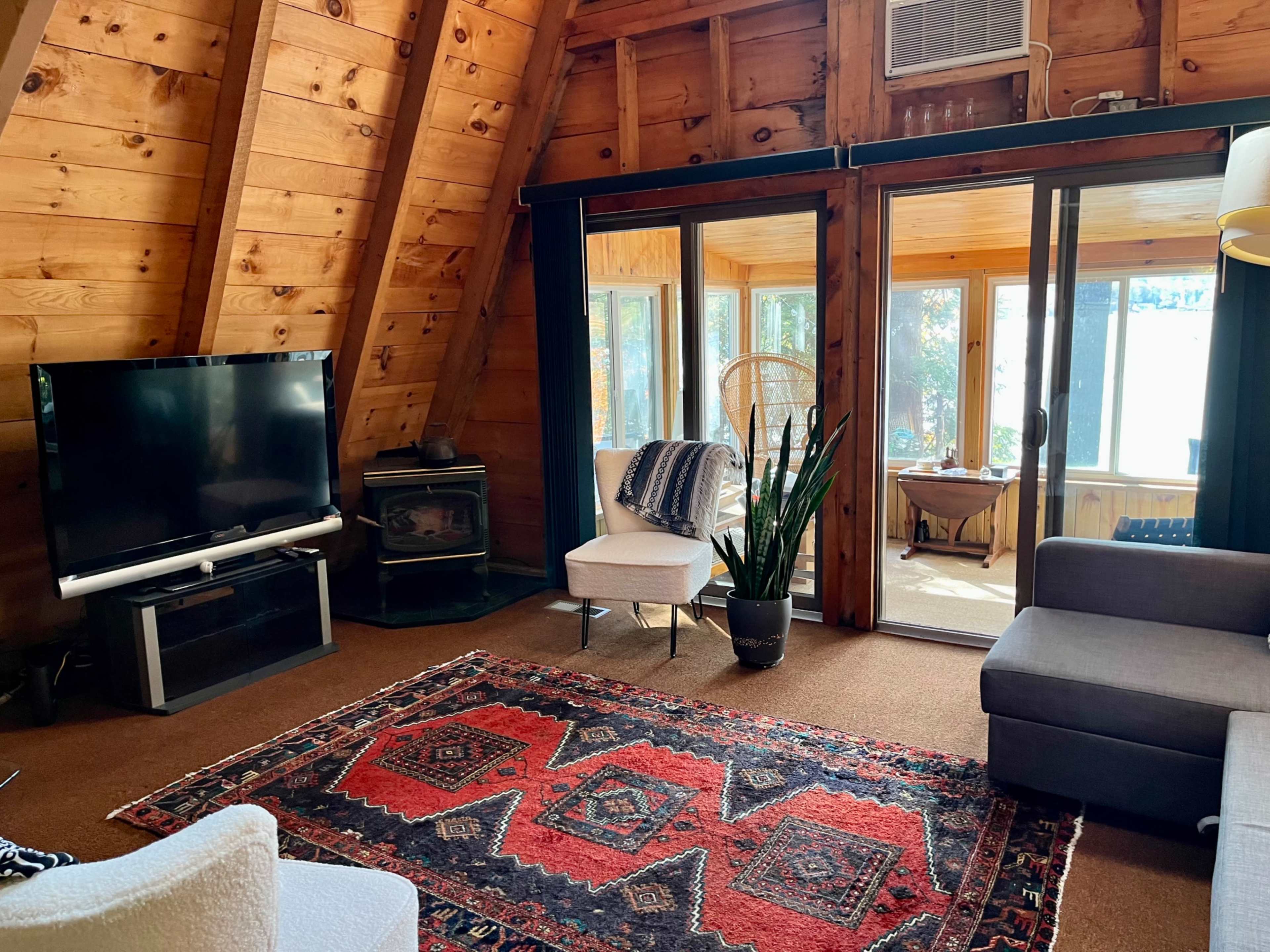 The image shows a wood-paneled living room with a television, a fireplace, and large windows overlooking a sunroom.
