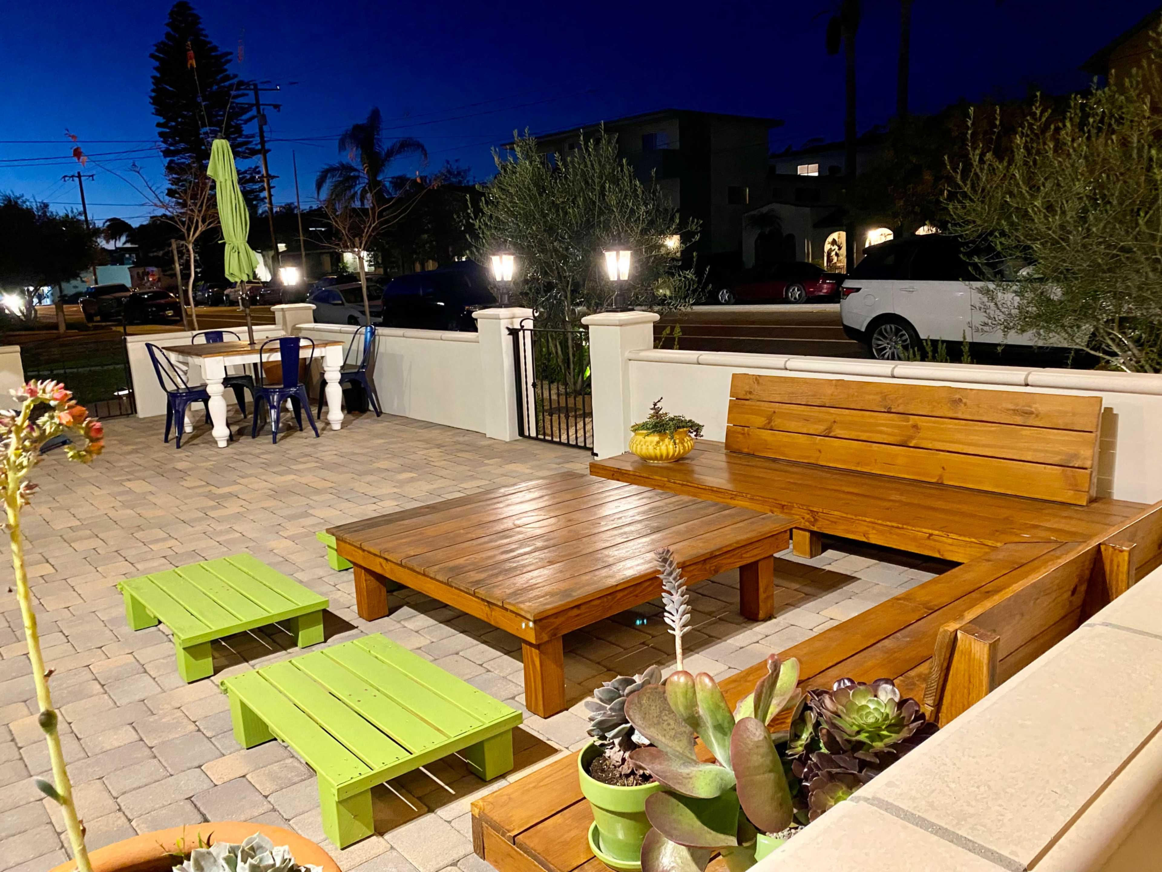 A patio area featuring wooden seating and a coffee table, surrounded by potted plants, under evening lighting.