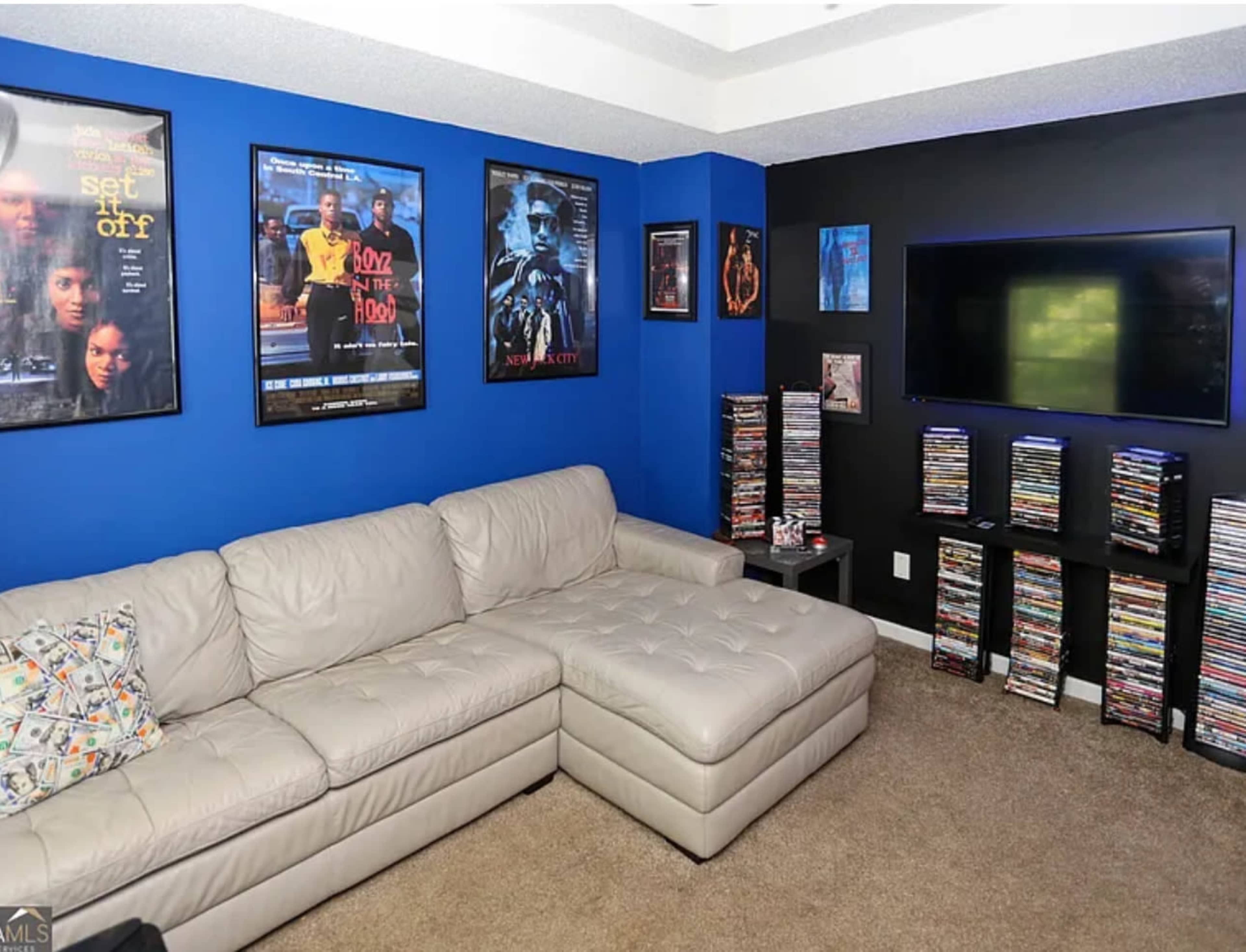 The room features a beige sectional sofa, a blue accent wall with movie posters, and a black wall with a mounted television and a collection of DVDs displayed on a shelf.