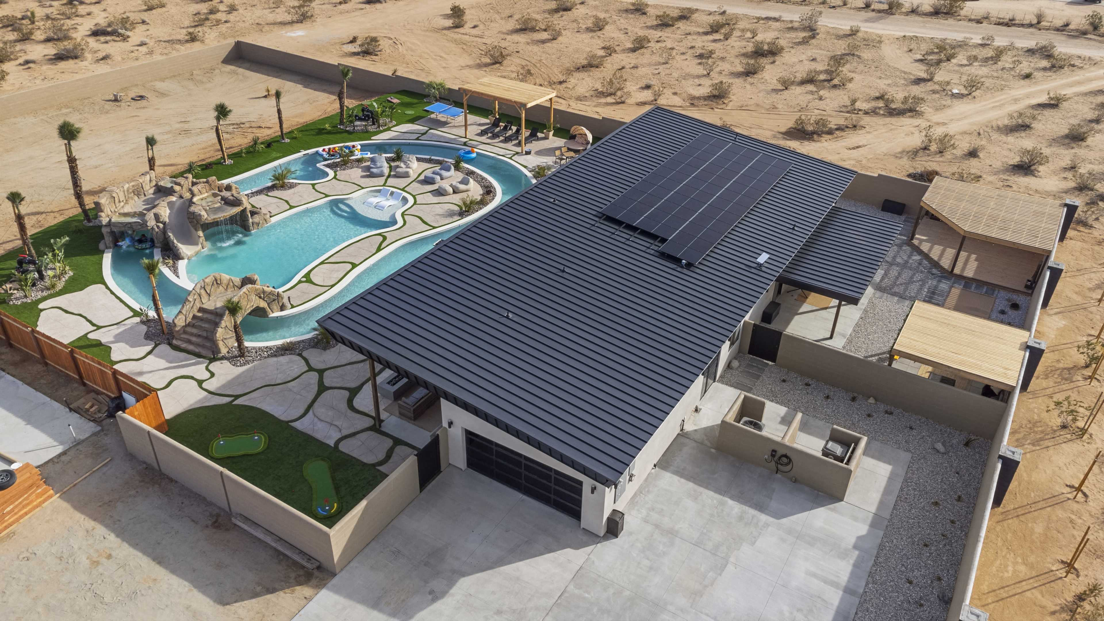 An aerial view of a modern house features a large swimming pool with a unique shape and landscaping, surrounded by palm trees and outdoor amenities in a desert setting.