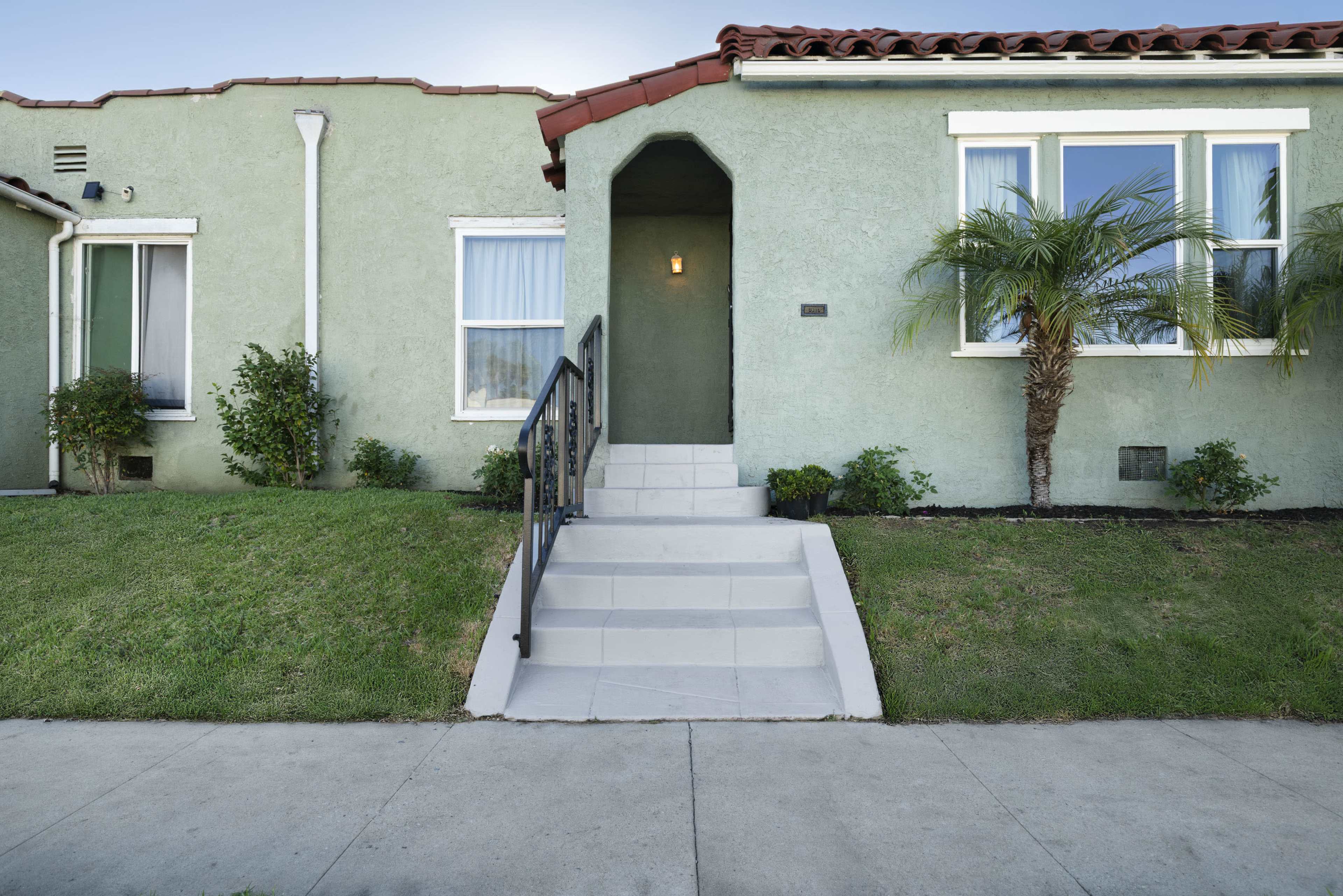 The image shows a green stucco house with a red tile roof, featuring a short set of stairs leading to a front door and two small palm trees in the yard.