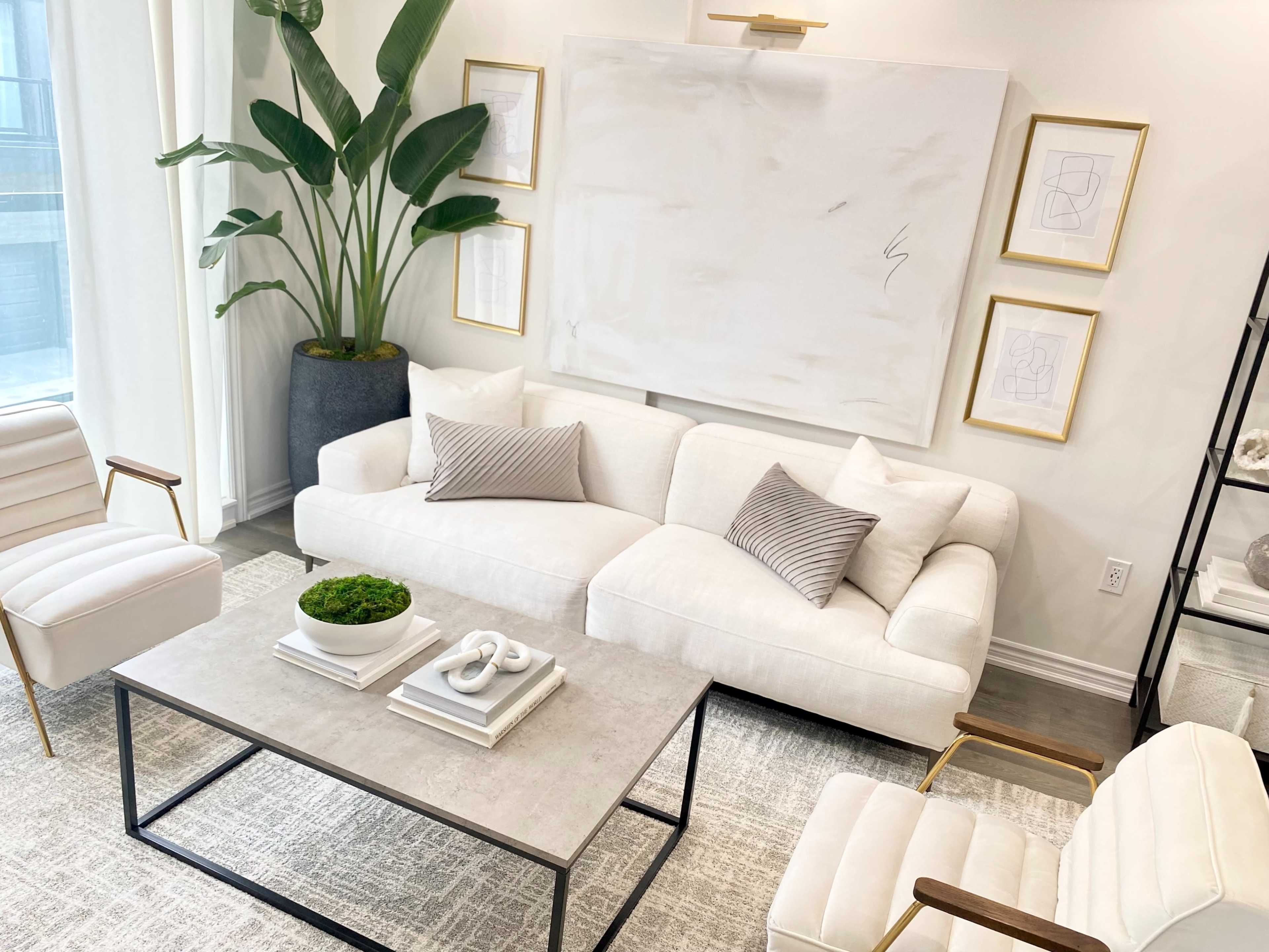 A modern living room features a light-colored sofa with decorative pillows, a minimalist coffee table, and framed artwork on the walls, alongside large green plants.