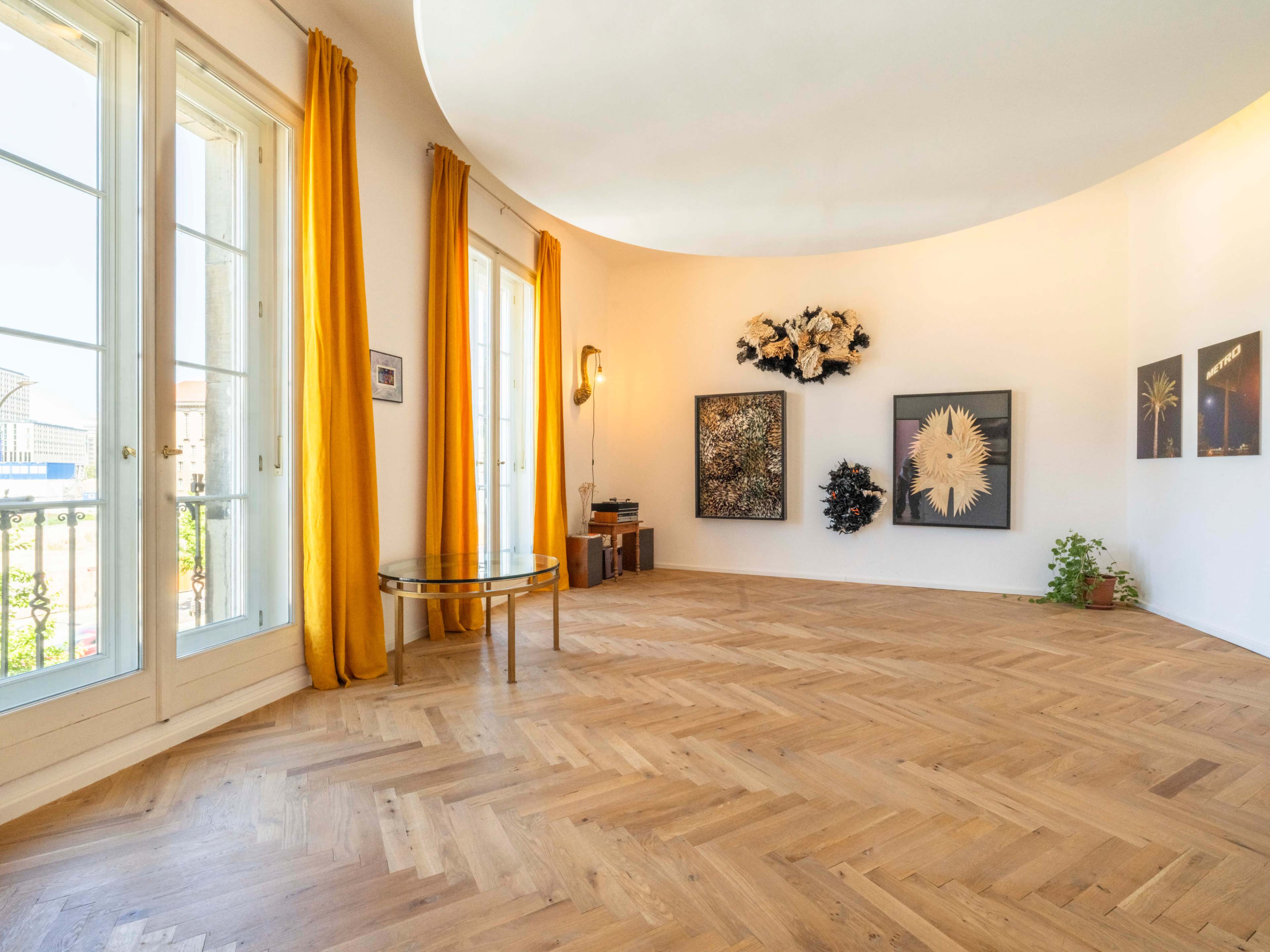 The room features large windows with curtains, hardwood flooring arranged in a herringbone pattern, and several artworks displayed on the walls.