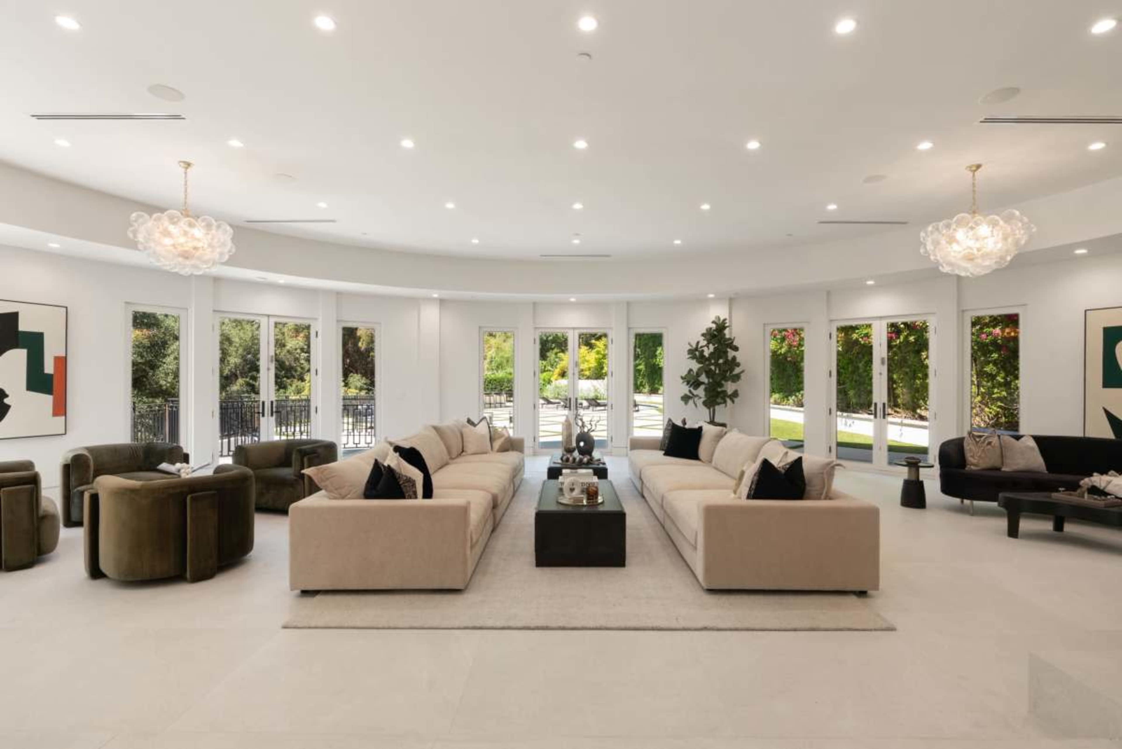 The image shows a spacious, modern living room with large windows, two cream-colored sofas facing each other, and minimalist decor including artwork and coffee tables.