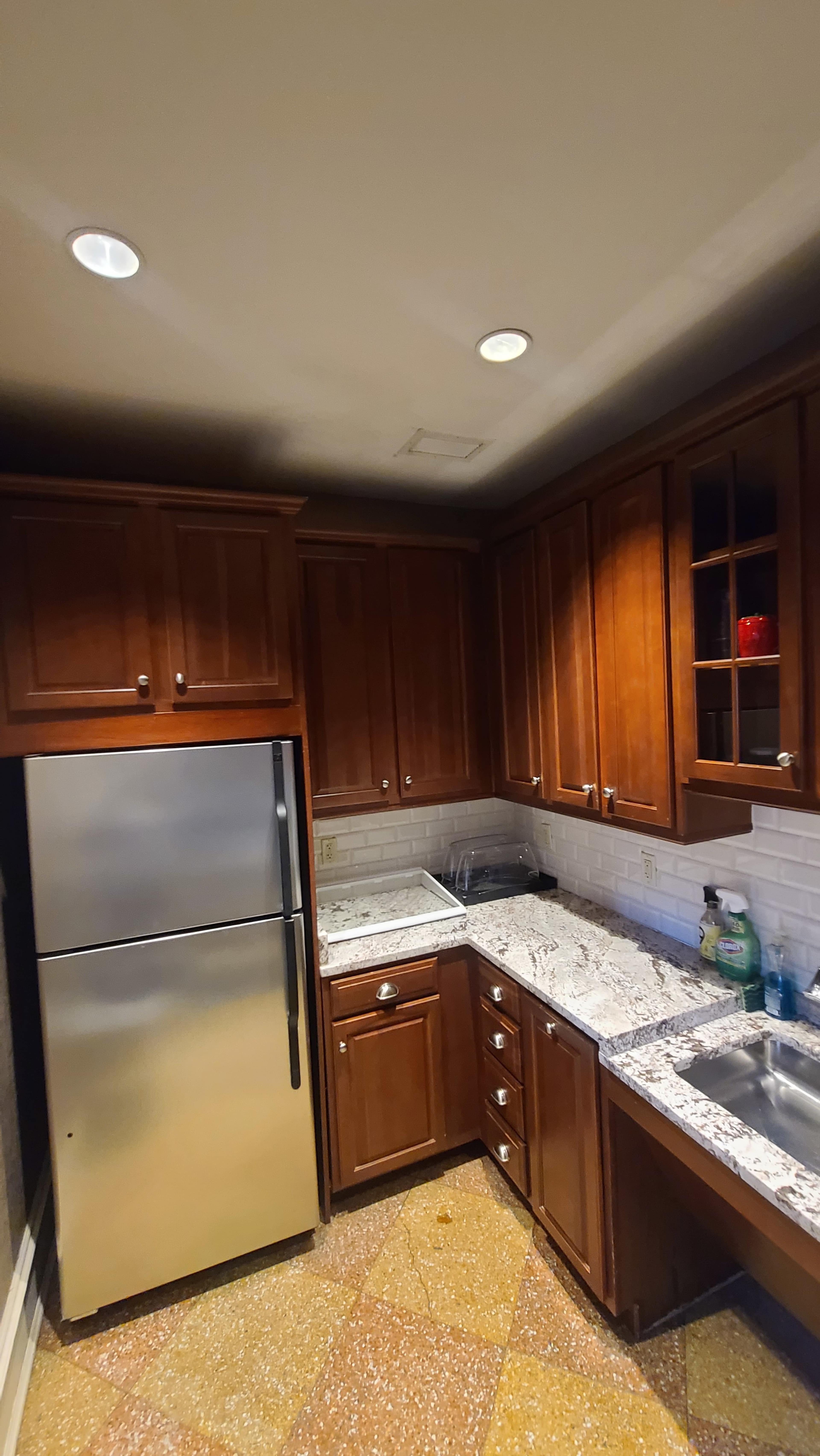 The image shows a small kitchen featuring dark wood cabinetry, a stainless steel refrigerator, and a countertop with a speckled granite surface.